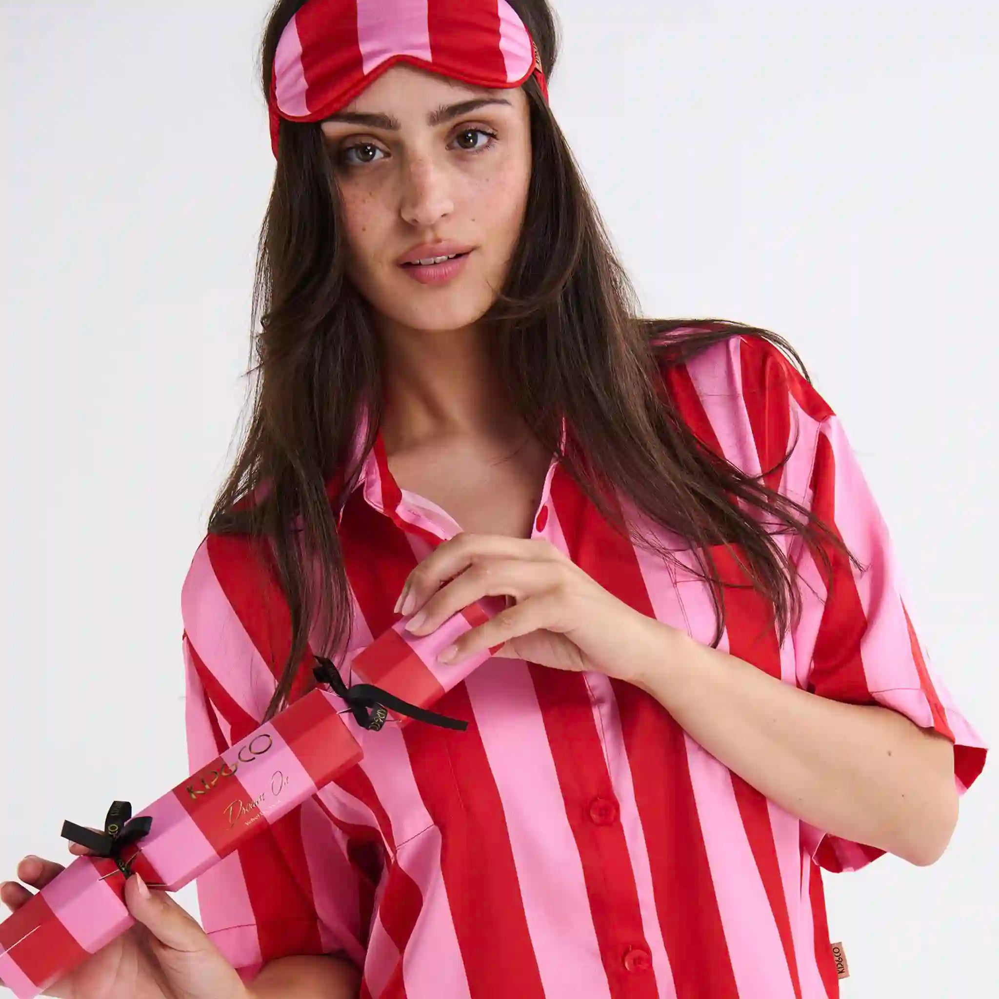 Woman wearing a red and pink striped pajama set with matching sleep mask on a white background