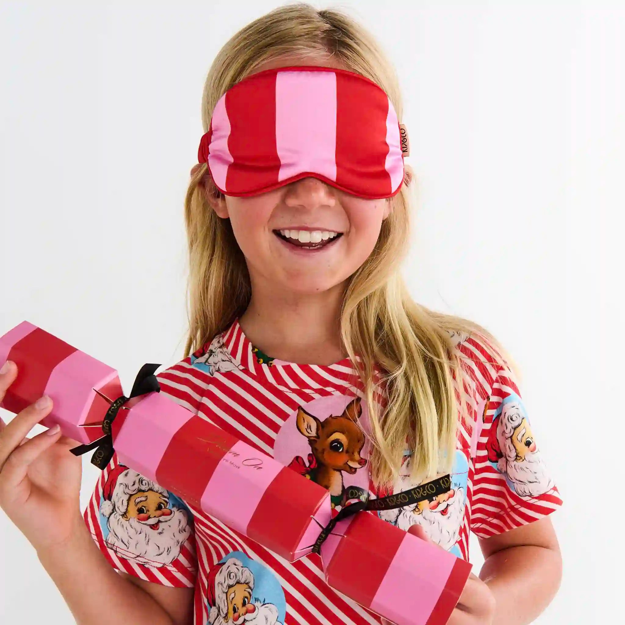 Child wearing a red and pink striped eye mask and holding a large pink and red cracker.