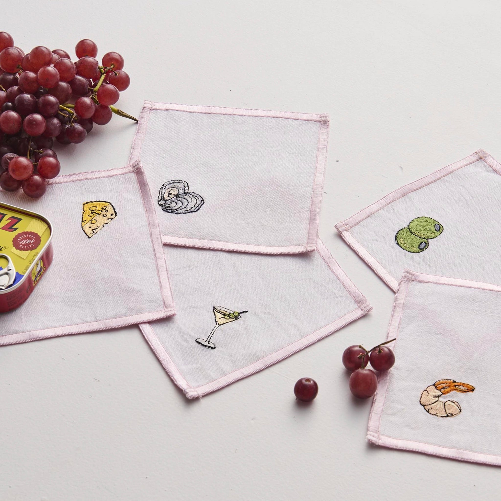 Set of embroidered napkins with various designs on a light surface with grapes and a can.