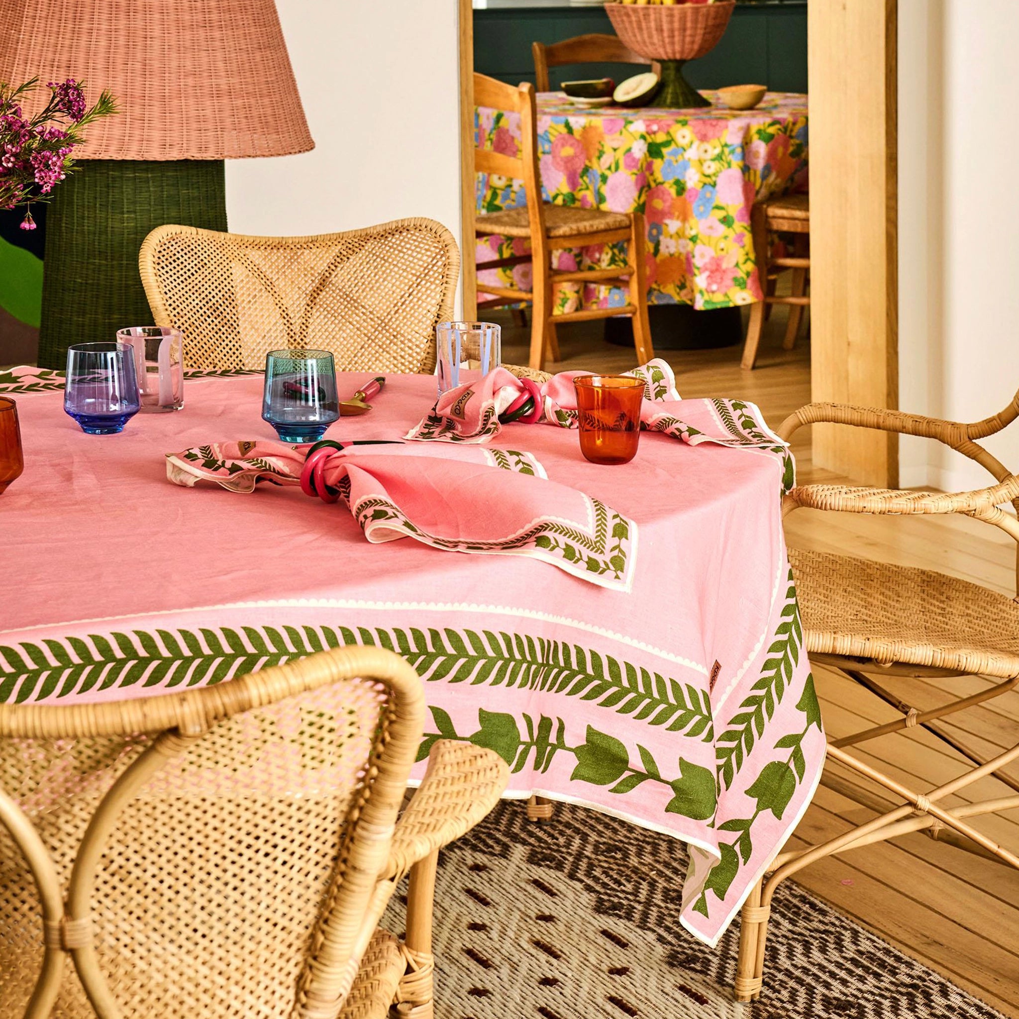 Dining room with a table set for a meal, featuring a pink tablecloth with green leaf patterns and wicker chairs.