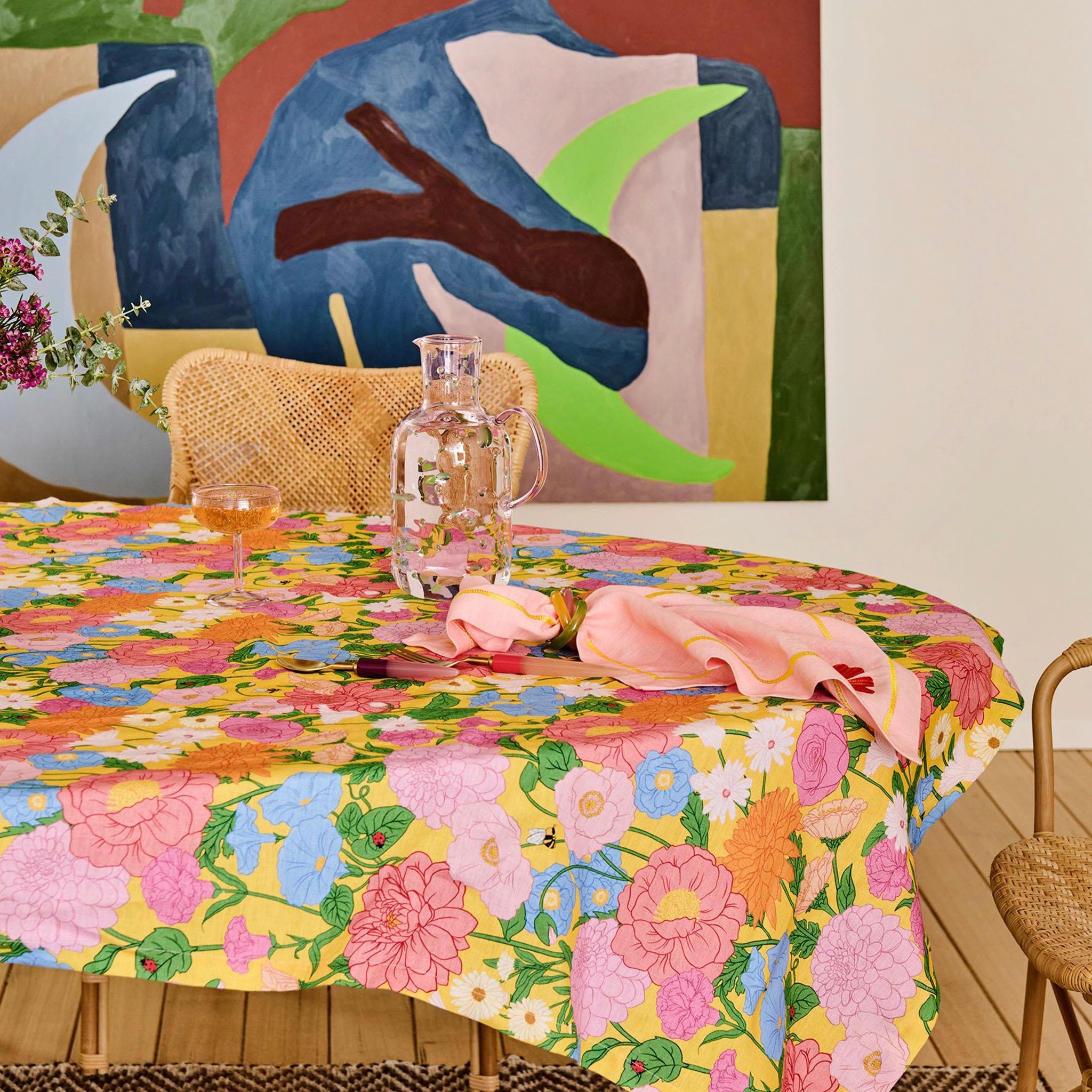 Dining table with a colorful floral tablecloth, glasses, and a pink napkin, with a abstract painting in the background.