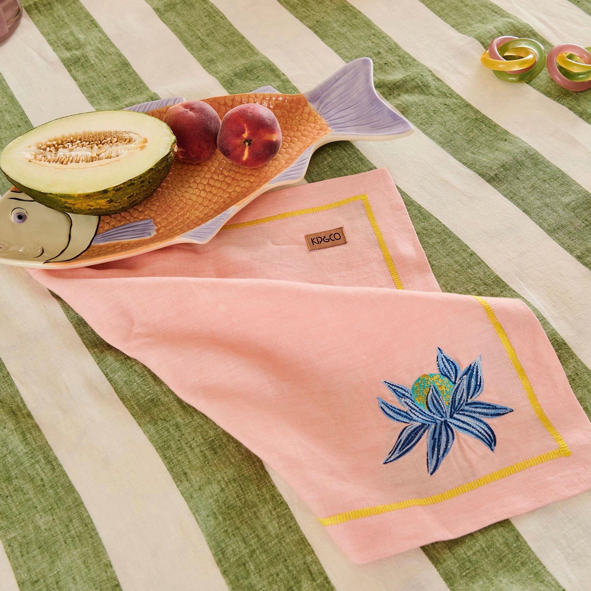 Pink towel with floral design on a green and white striped tablecloth with fruit and fish-shaped plate.