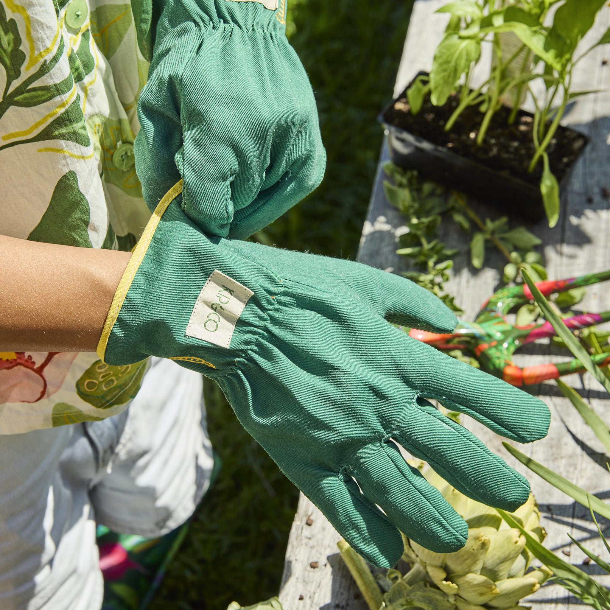Green gardening gloves held by a person with plants and gardening tools in the background
