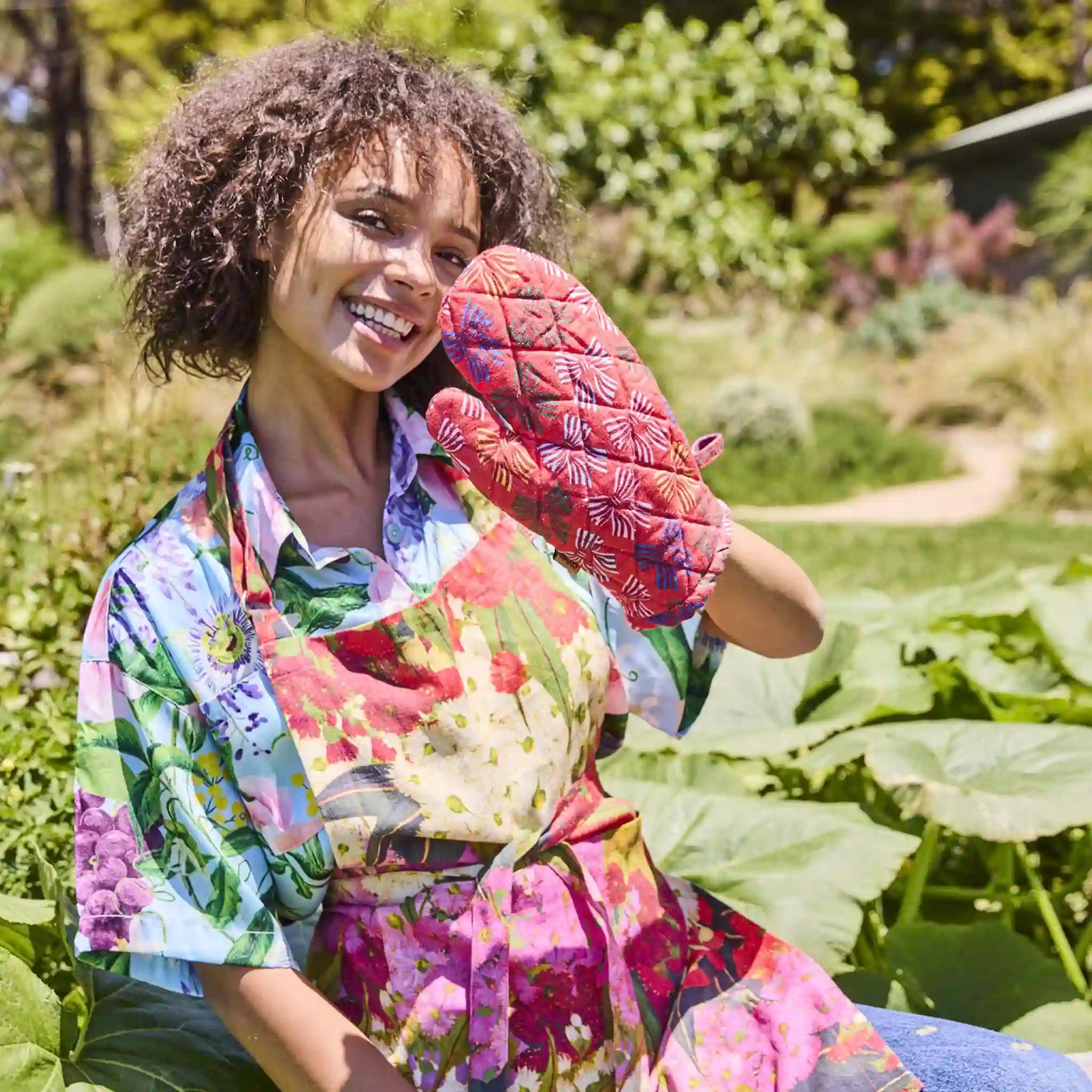 Woman in a garden wearing a colorful apron and oven mitt, holding a leafy green vegetable.