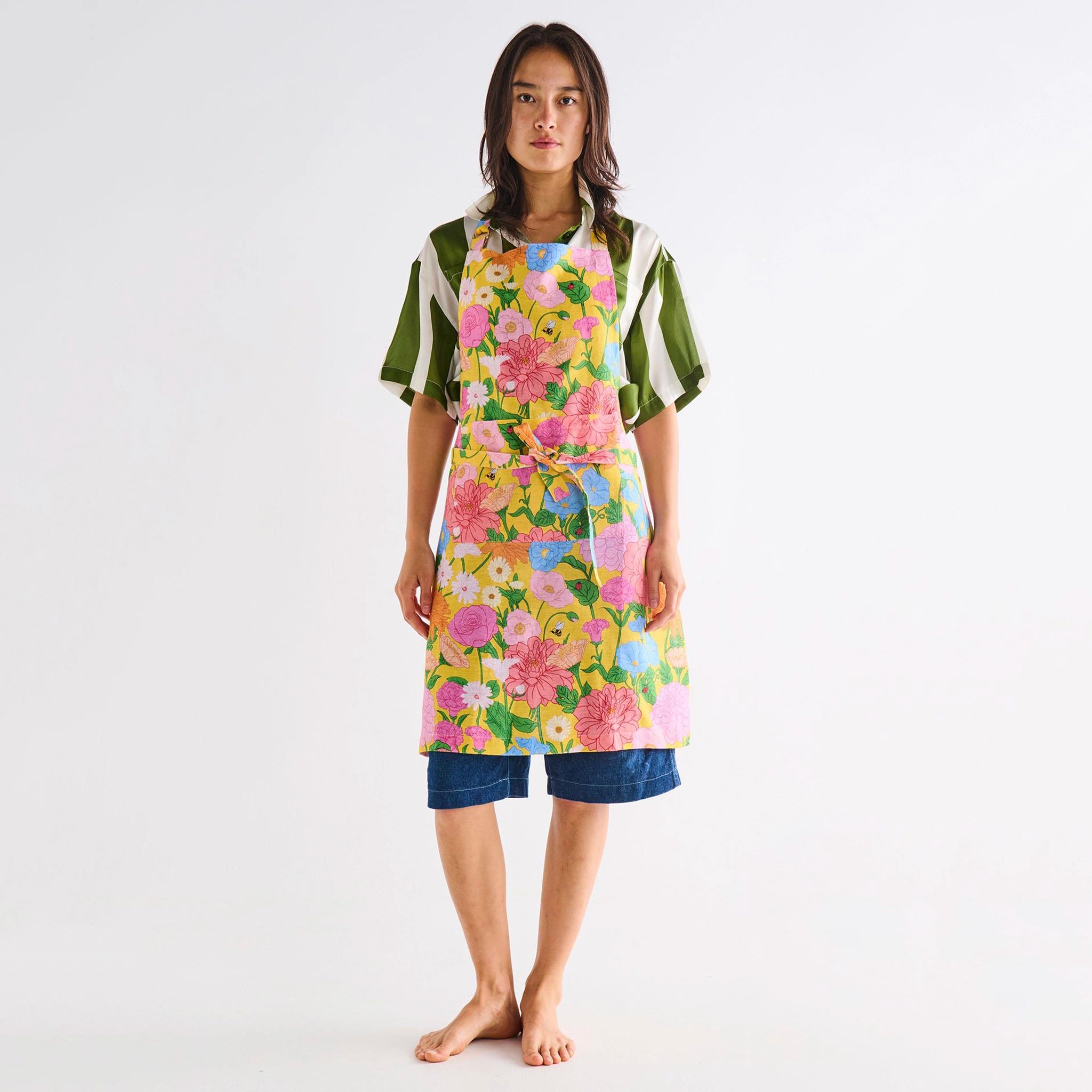 Woman wearing a colorful floral apron over a green shirt and blue shorts on a white background
