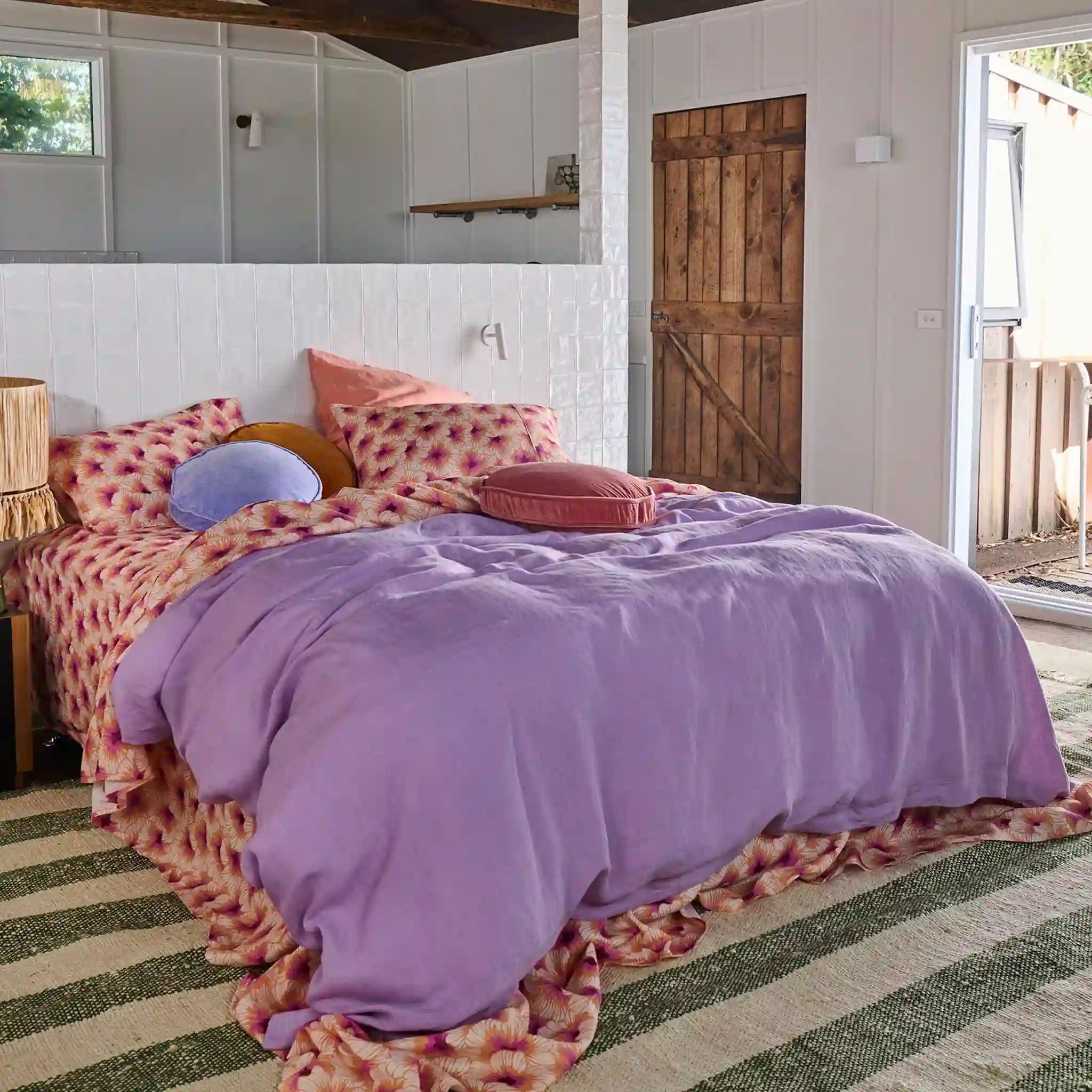 Bedroom with a bed covered in a purple comforter and patterned bedding, wooden door, and striped rug.