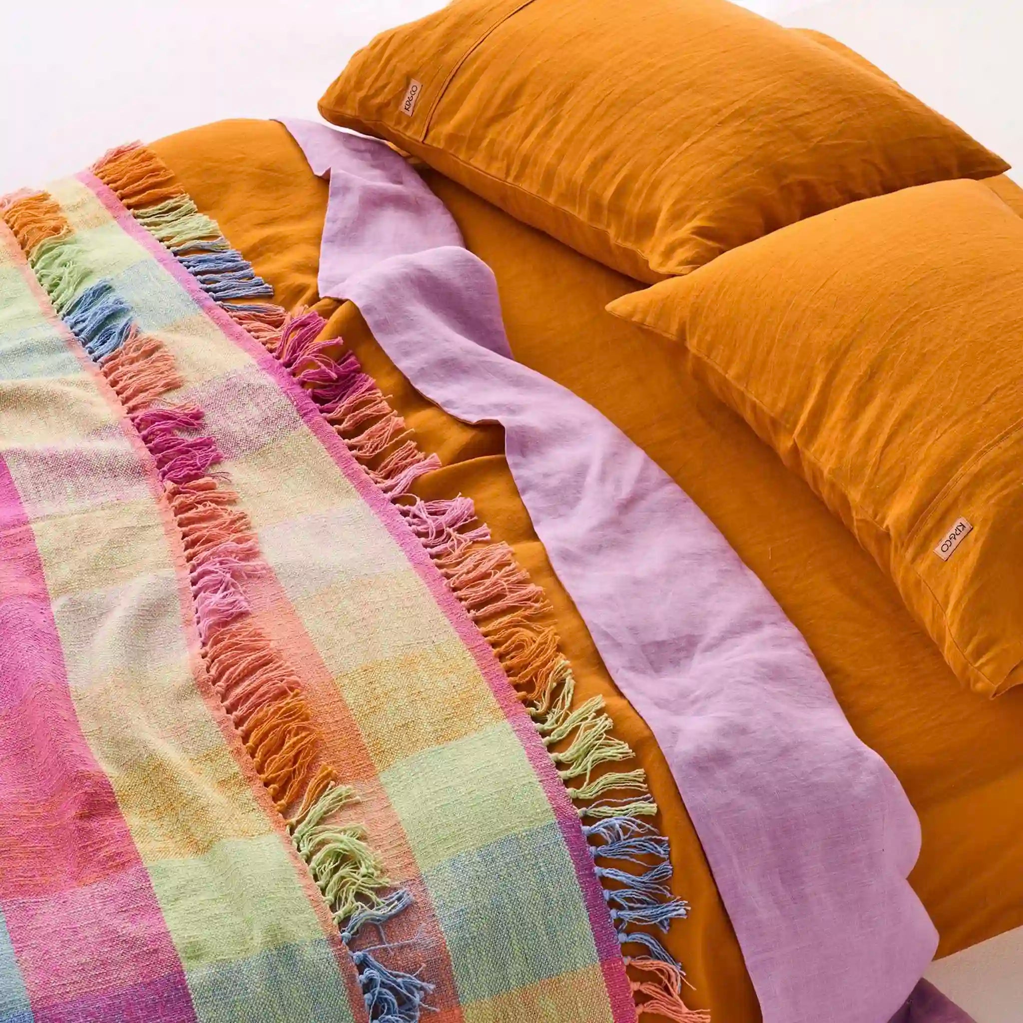 Multicolored plaid blanket with fringe on a bed with orange bedding and pillows.