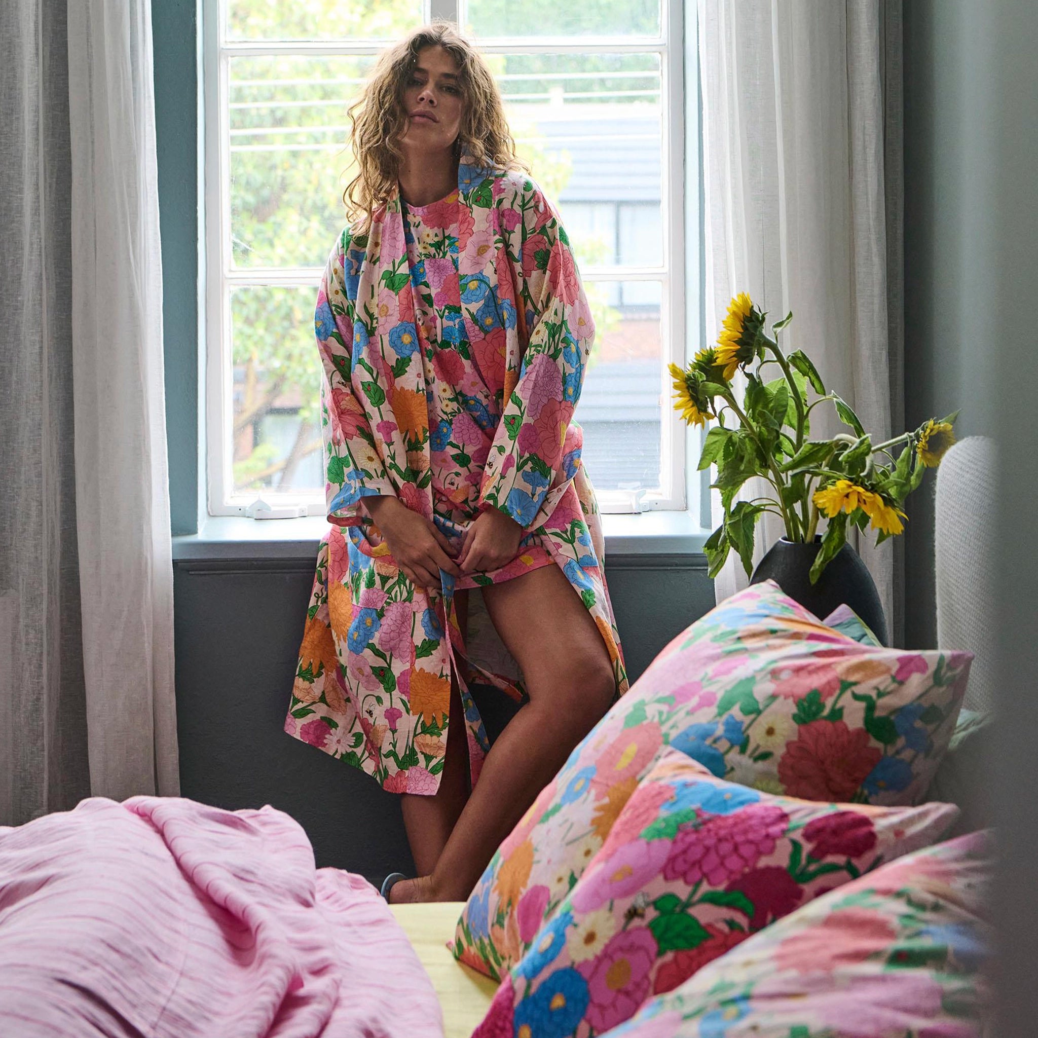 Woman in a colorful floral robe standing in a bedroom with matching bedding and pillows.