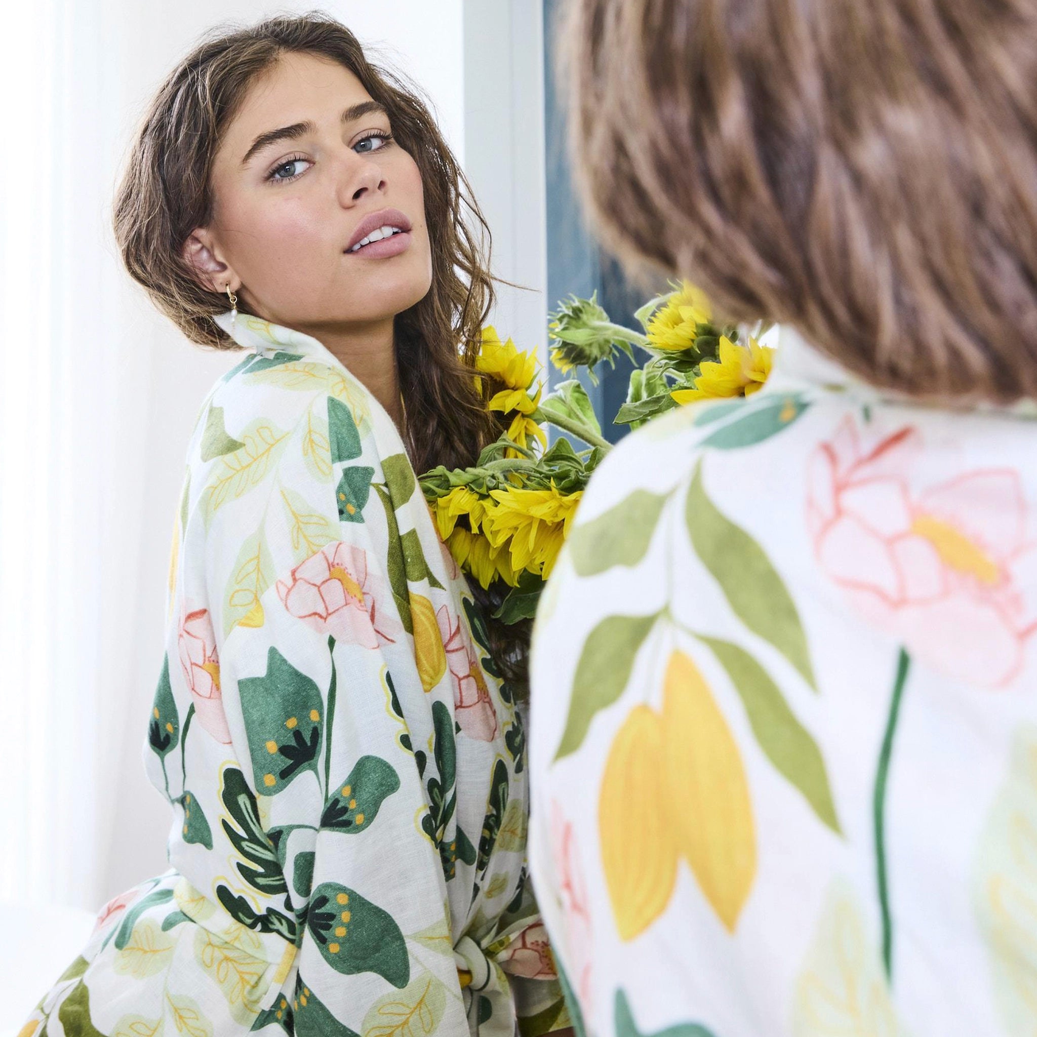 Woman wearing a floral robe holding flowers, looking at herself in the mirror.