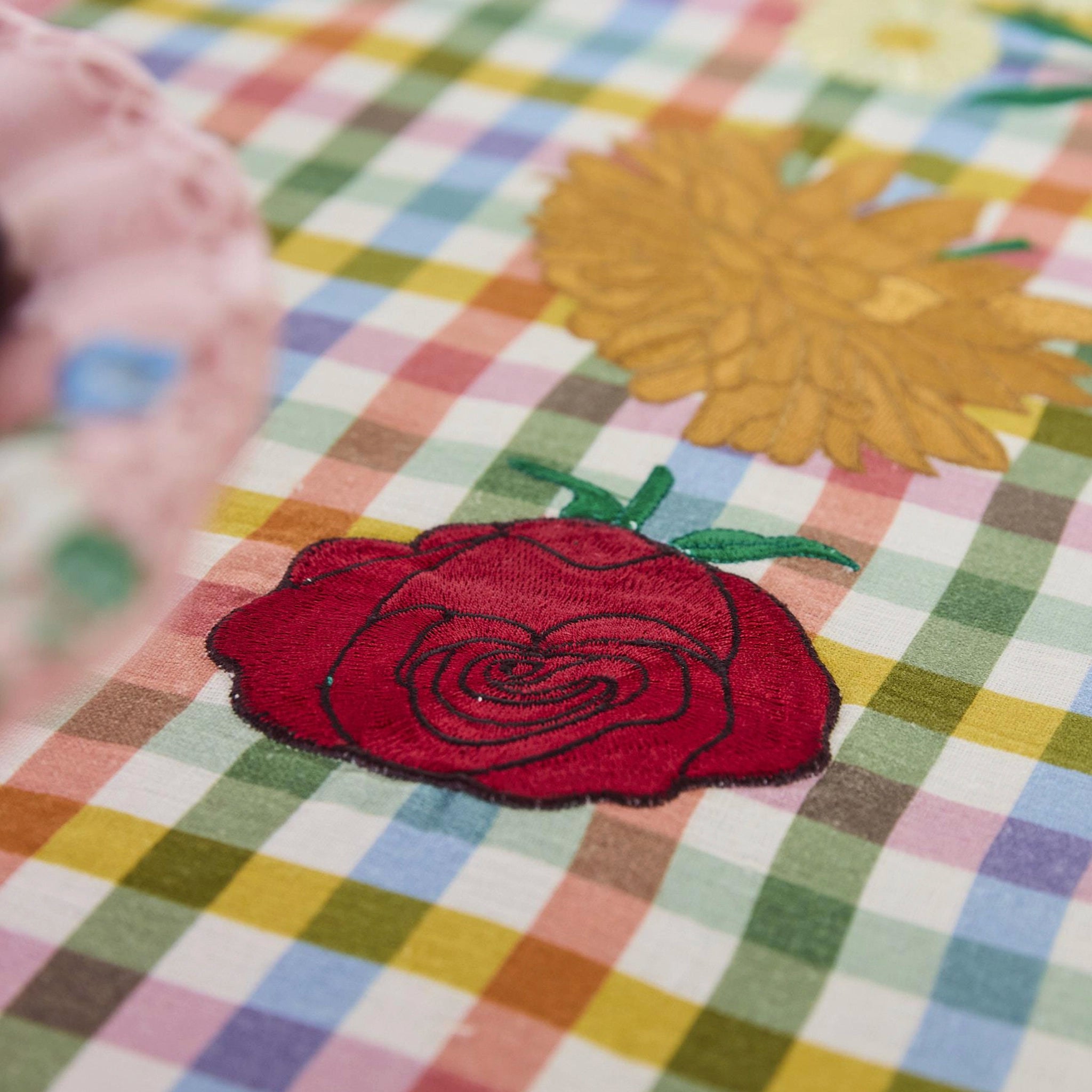 Embroidered red rose on a colorful checkered fabric with a yellow flower in the background.