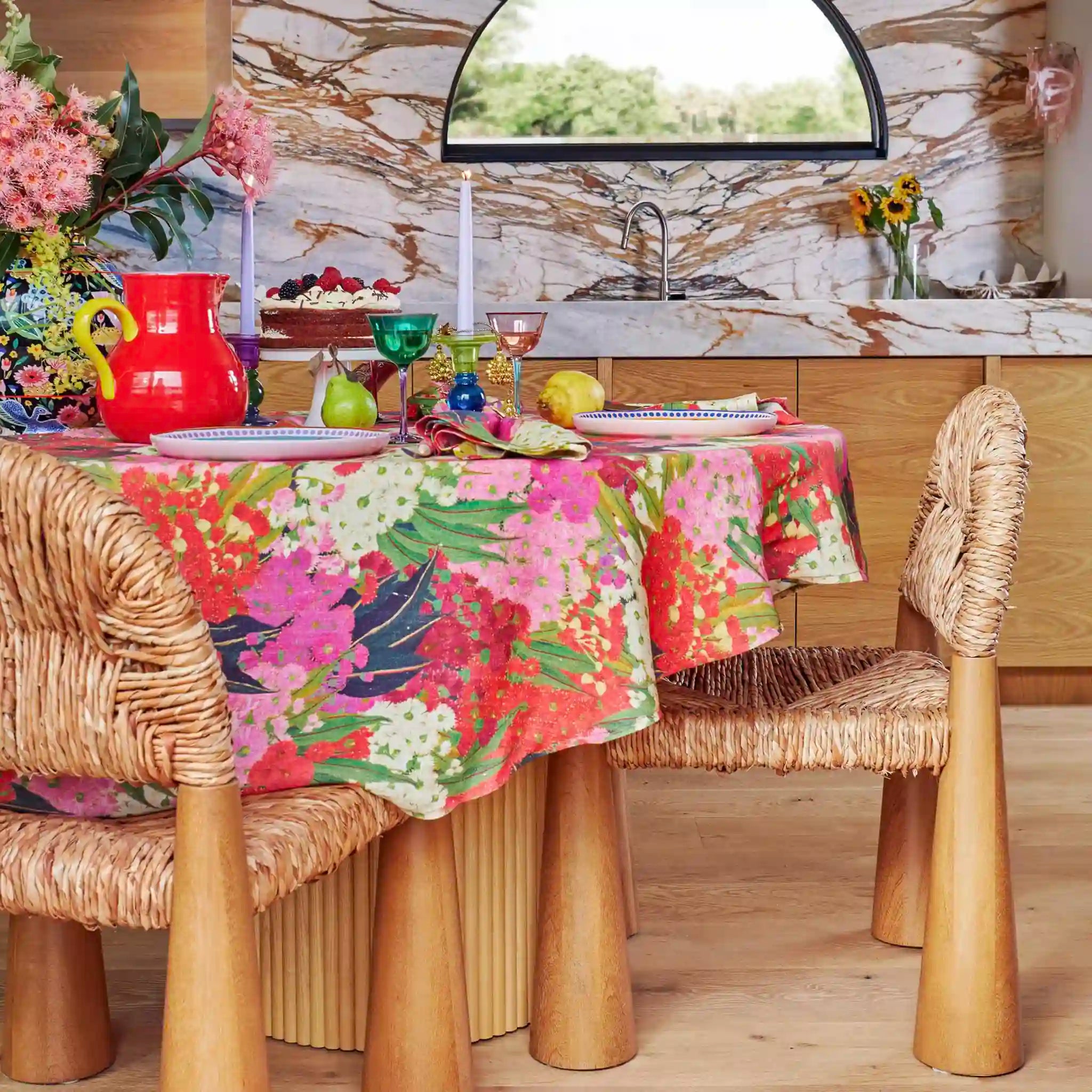 Dining table with a floral tablecloth, wicker chairs, and a kitchen in the background.