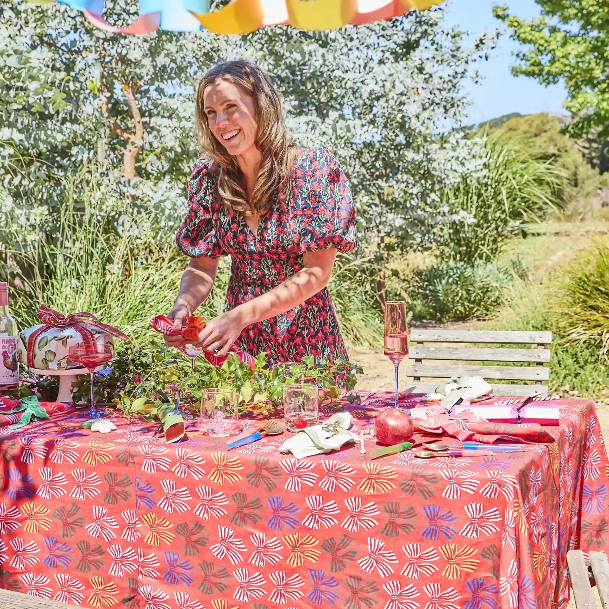 Woman setting a table outdoors with a floral tablecloth and decorations.