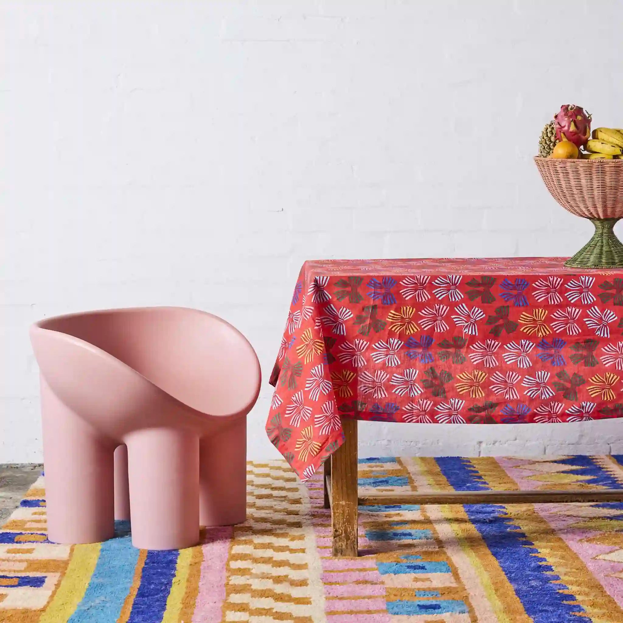Pink chair next to a table with a colorful tablecloth and fruit basket on a patterned rug.