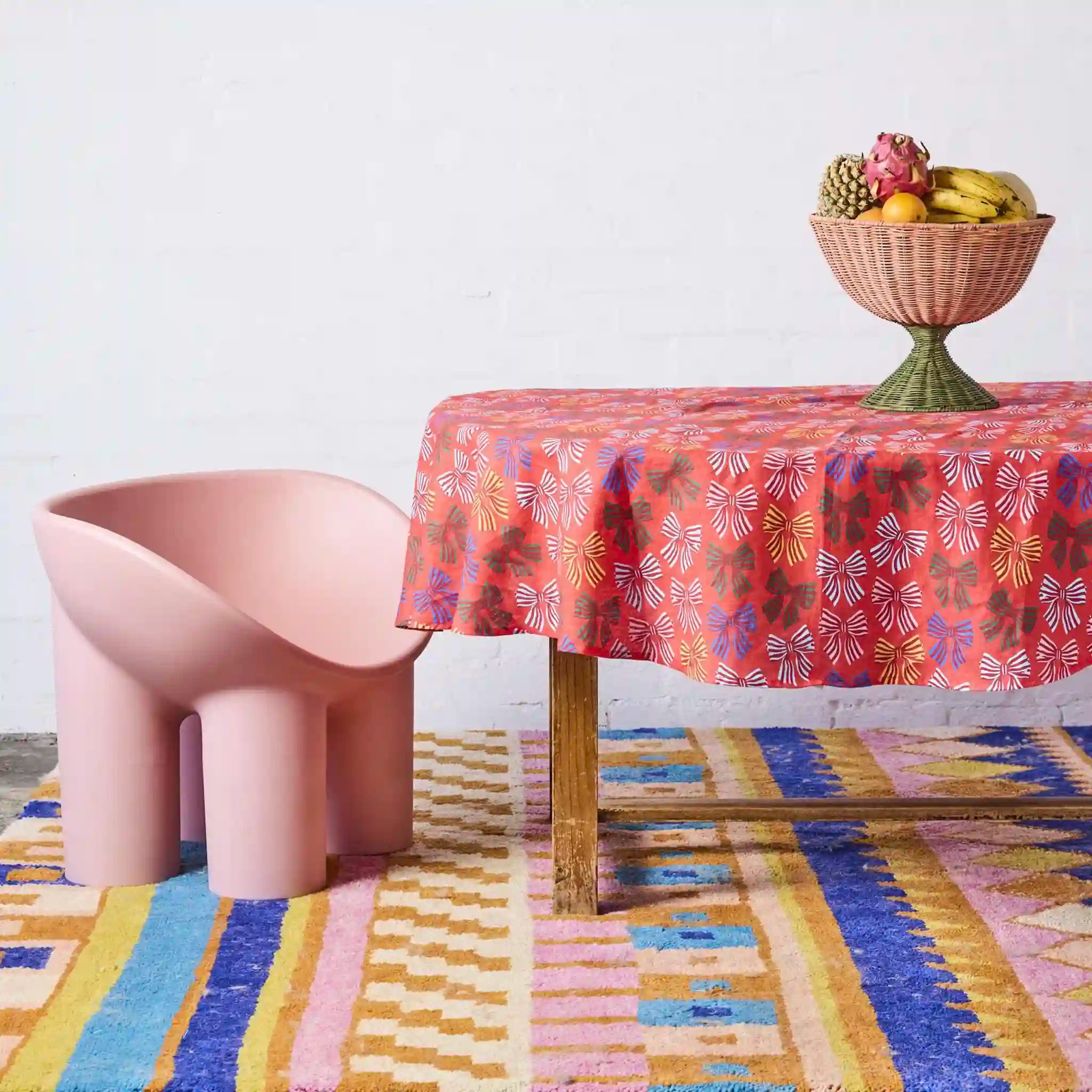 Table with a colorful tablecloth, pink chair, and fruit bowl on a patterned rug.