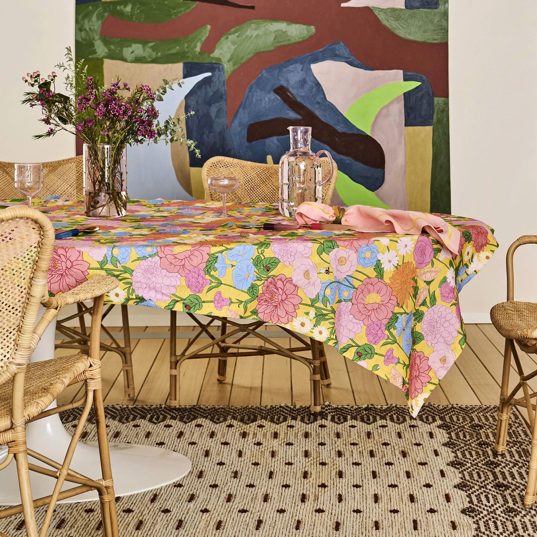 Dining table with a colorful floral tablecloth, surrounded by wicker chairs in a room with abstract art on the wall.