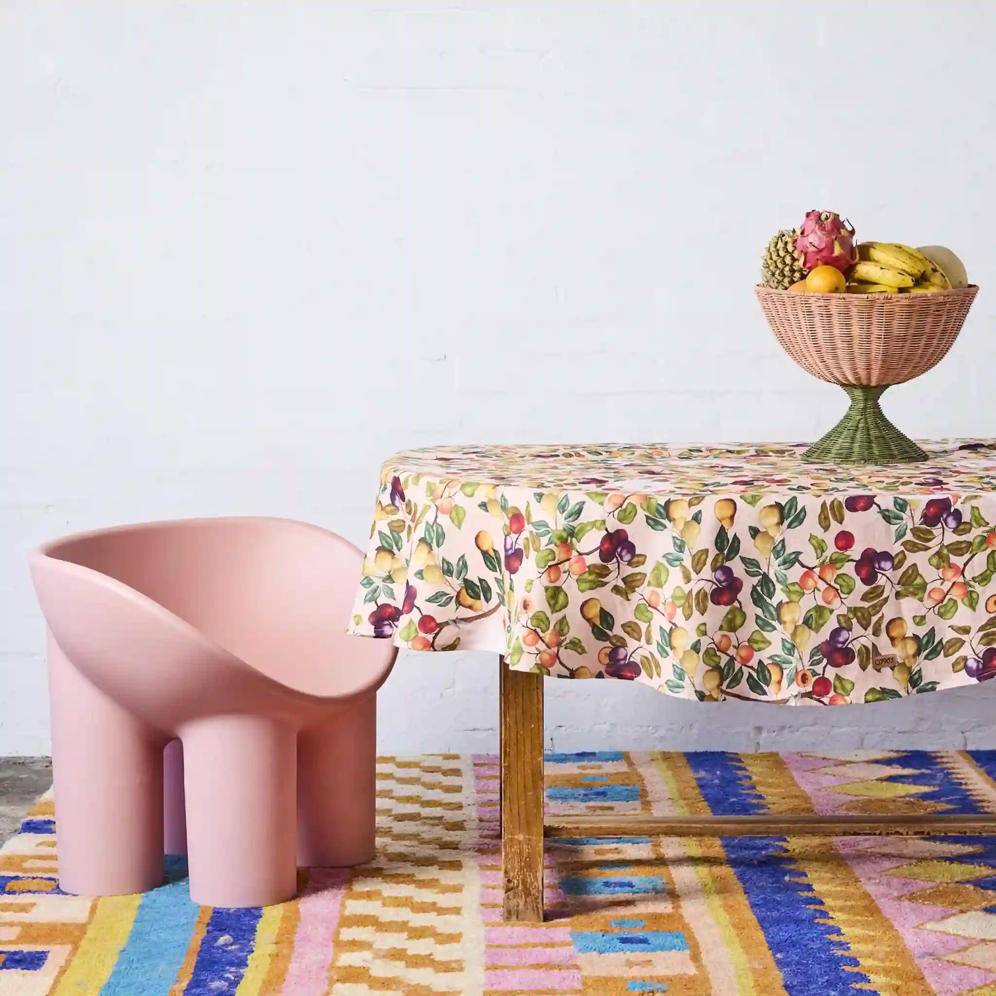 Table with a floral tablecloth, pink chair, and fruit bowl on a colorful rug.