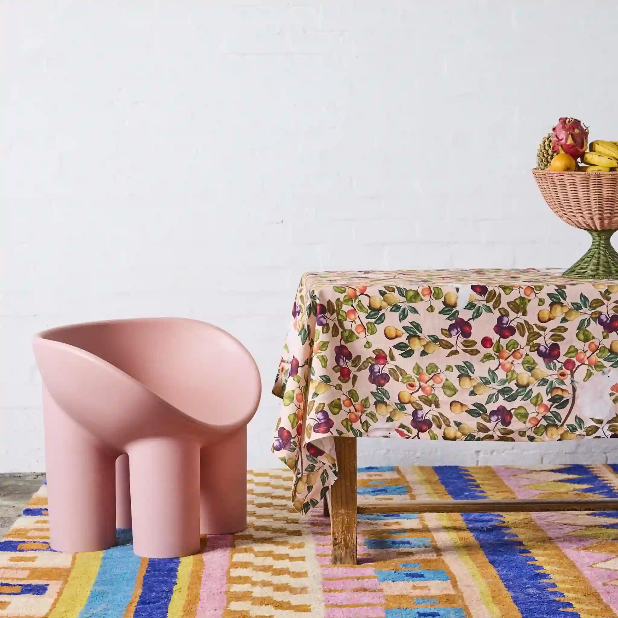 Pink chair next to a table with a floral tablecloth on a colorful rug.