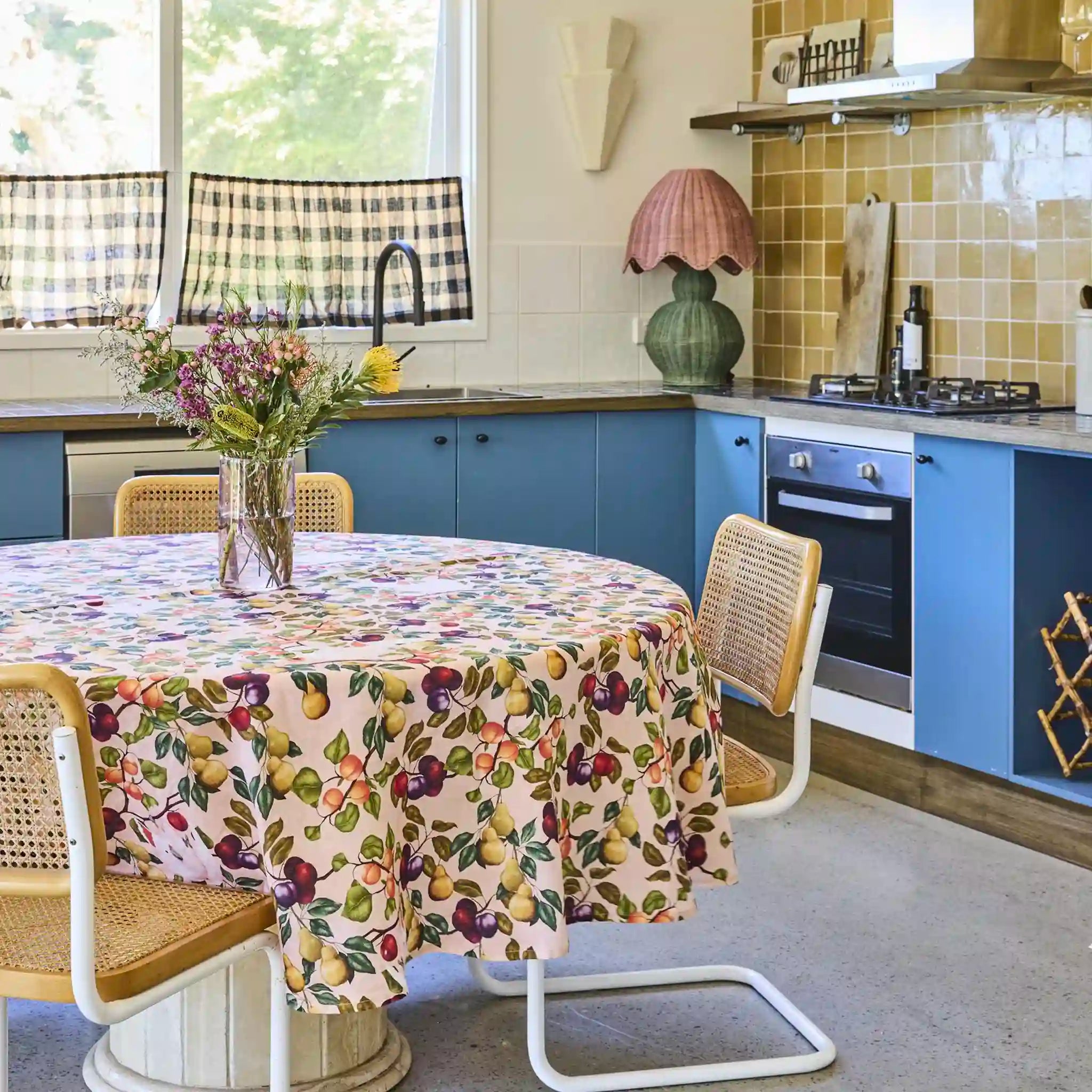 Kitchen with blue cabinets, round table with fruit-patterned tablecloth, and checkered curtains.