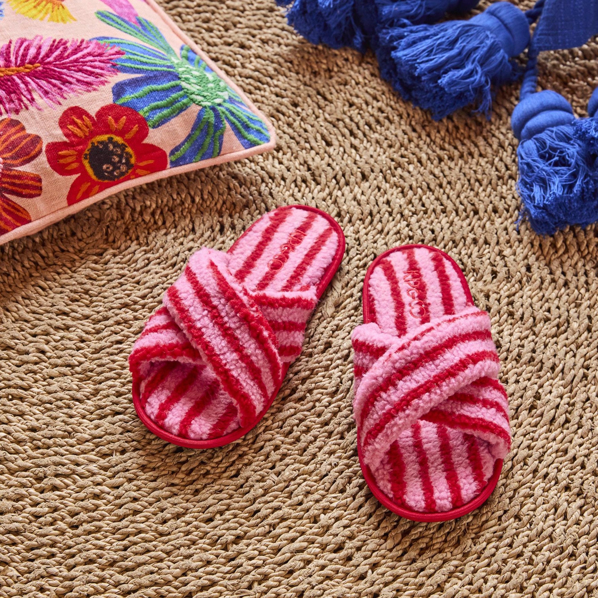 Red and pink striped slippers on a textured surface with a colorful pillow and blue decorative items in the background.