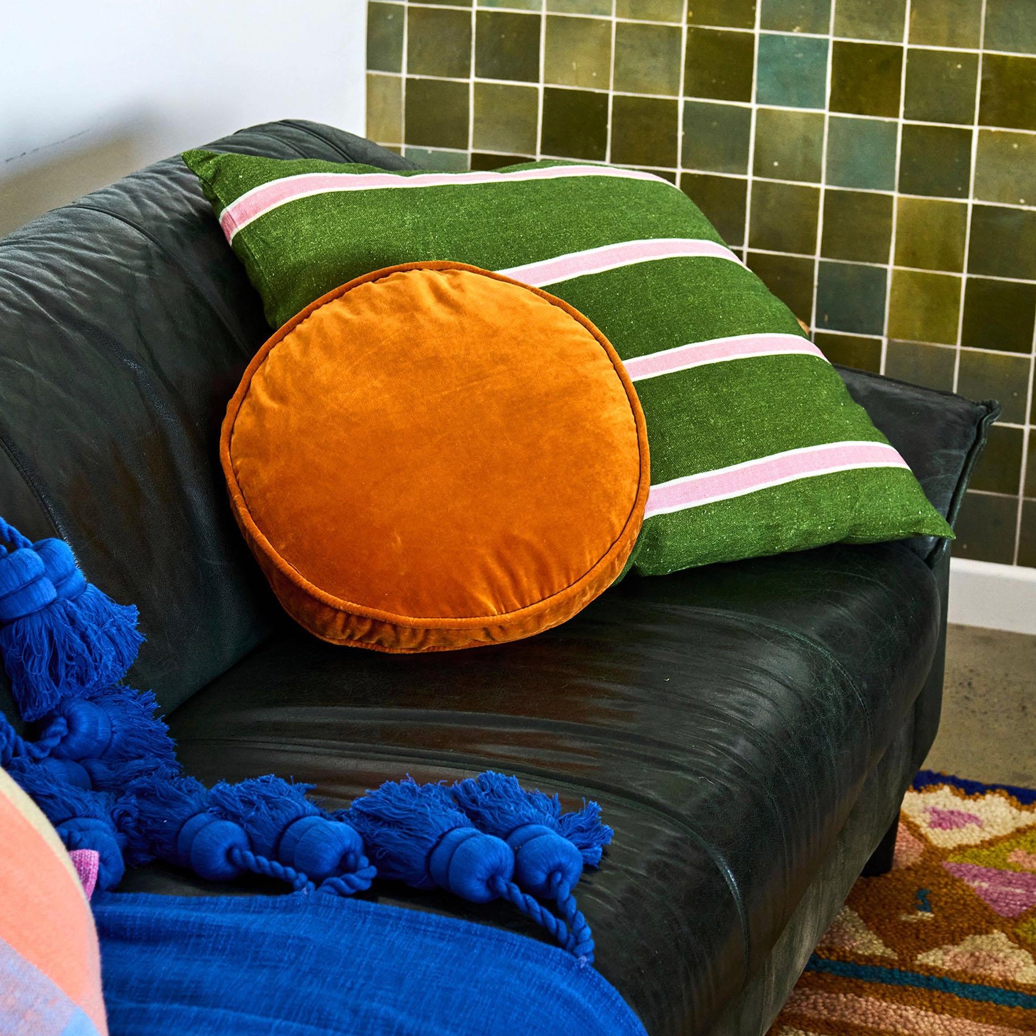Green cushion with pink stripes and an orange circle on a black couch against a green tiled wall.