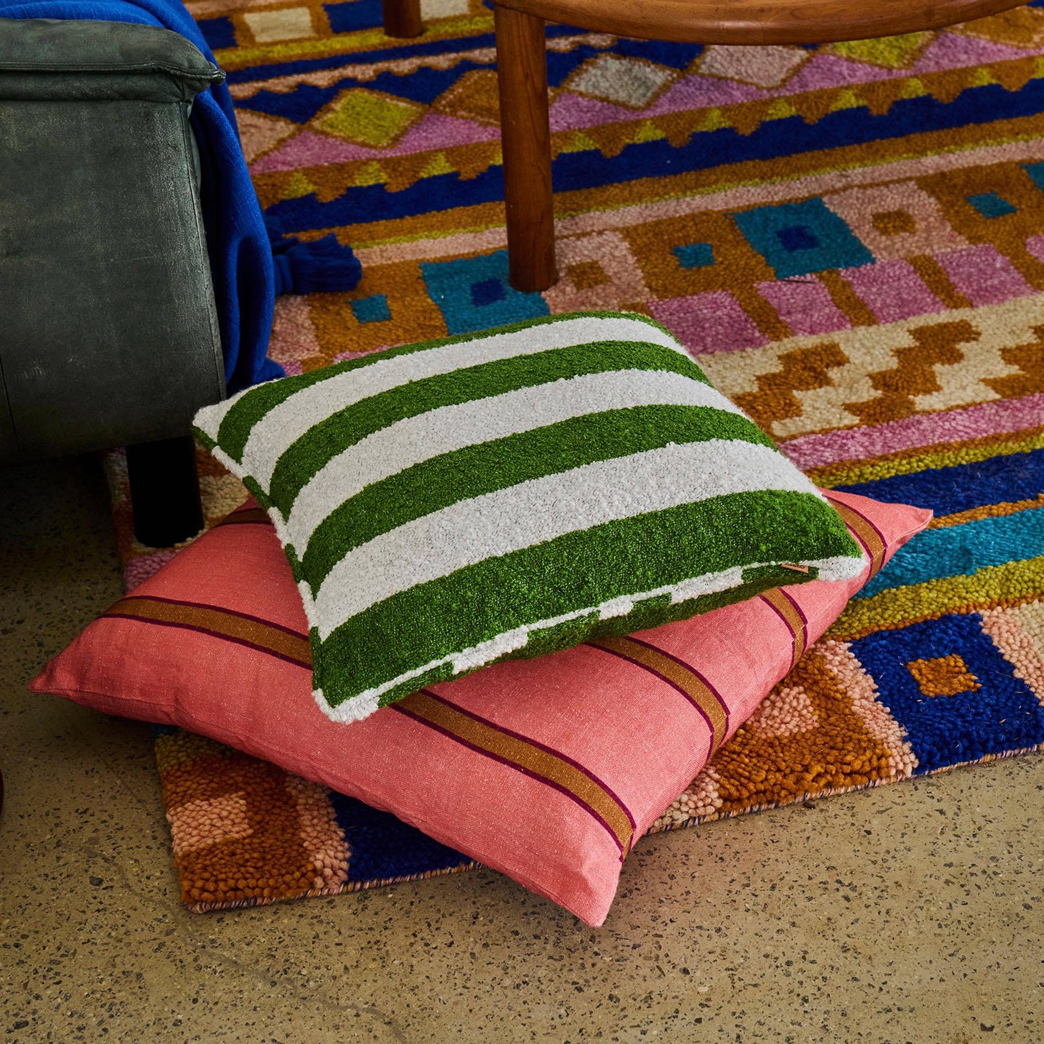 Green and white striped cushion on a pink cushion with a colorful geometric rug in the background.