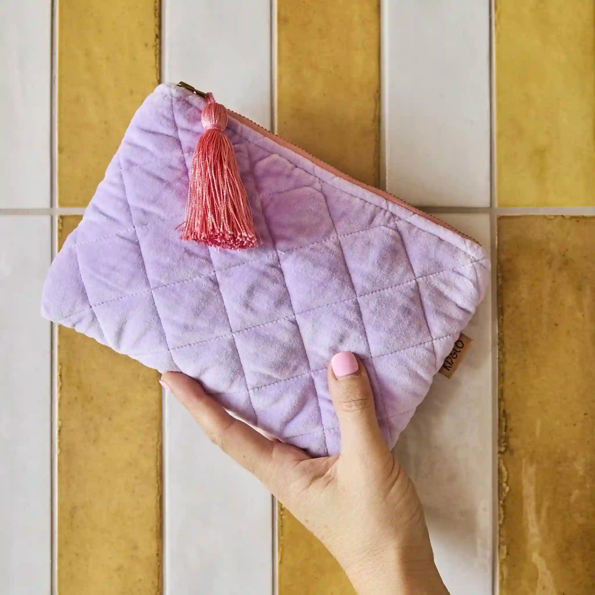 Purple quilted cosmetic bag with a pink tassel held against a tiled wall.