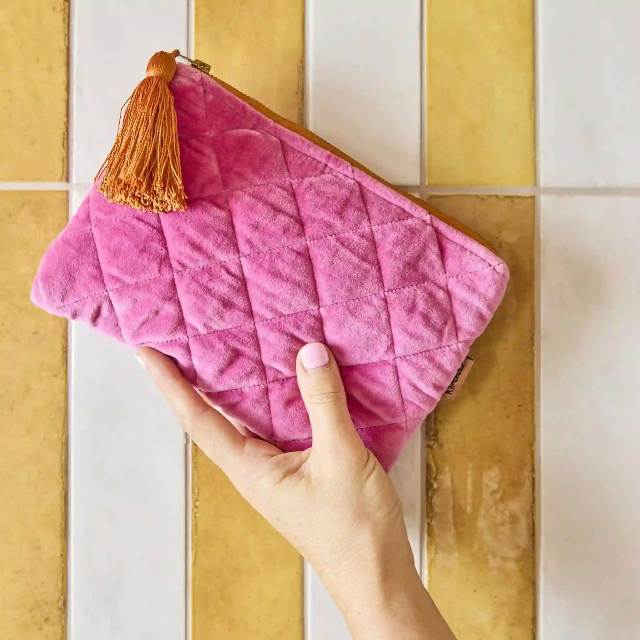 Pink quilted cosmetic bag with a tassel held by a hand against a tiled wall background
