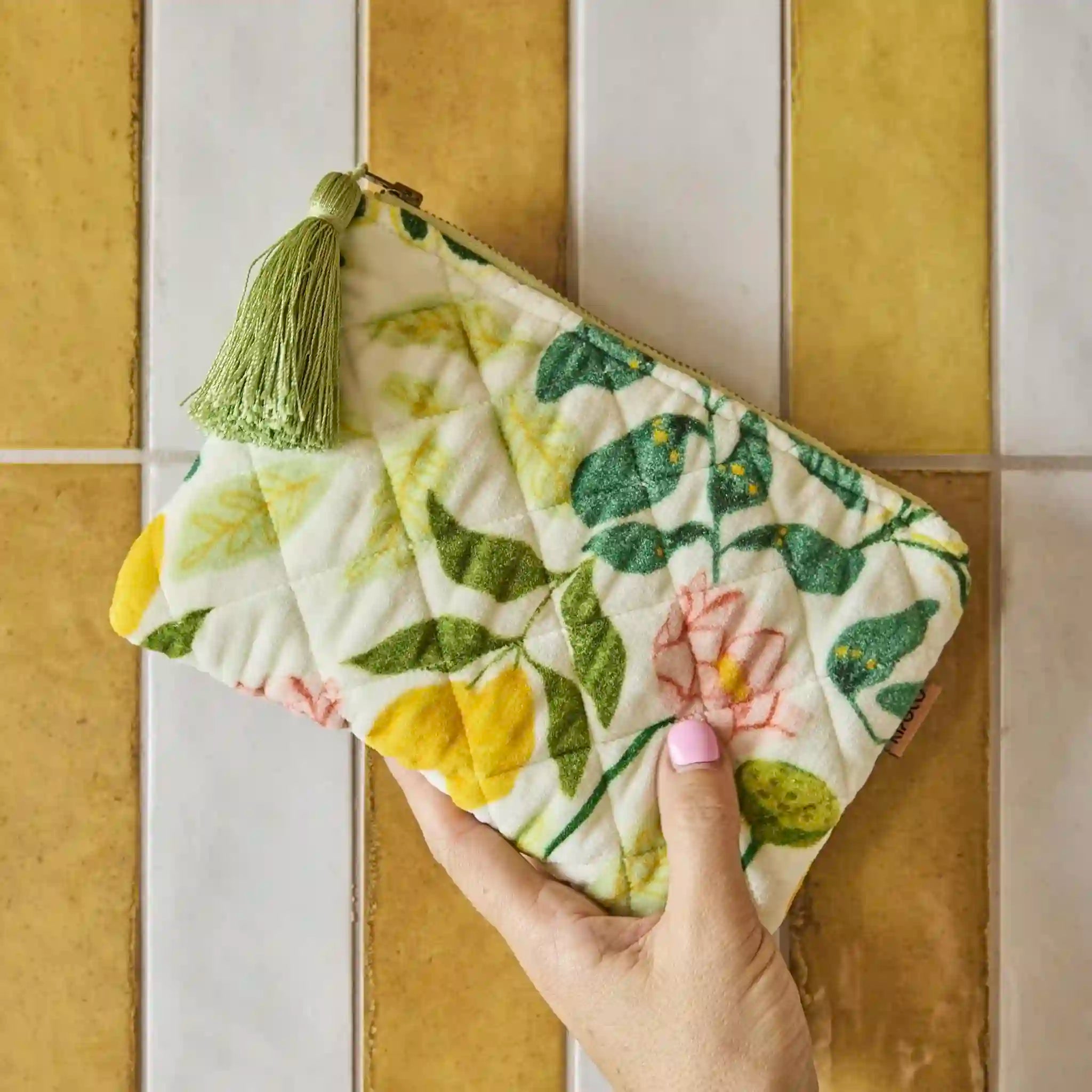 Hand holding a floral-patterned cosmetic bag with tassel against a tiled wall background