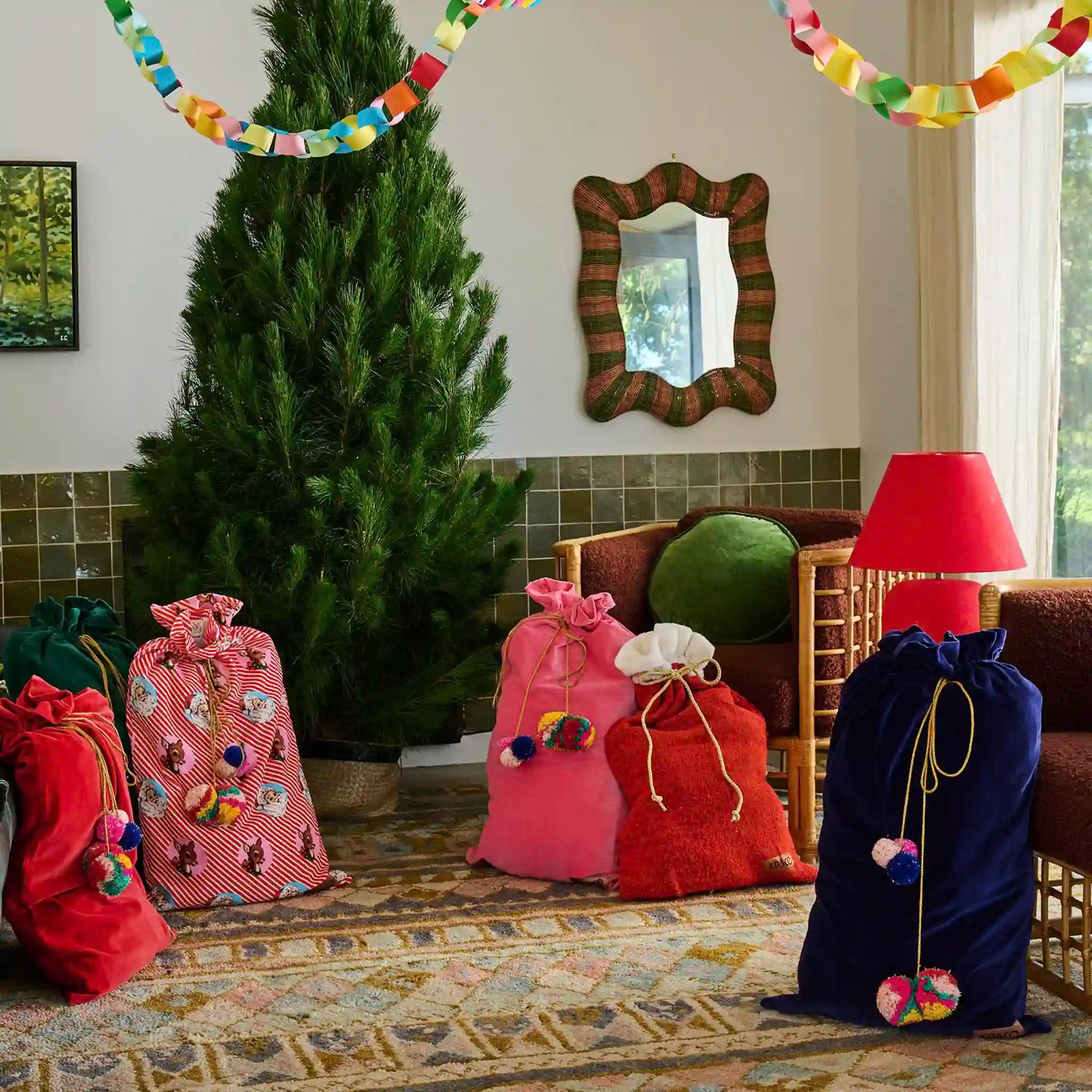 Decorative gift bags with colorful pom-poms in front of a Christmas tree in a living room.