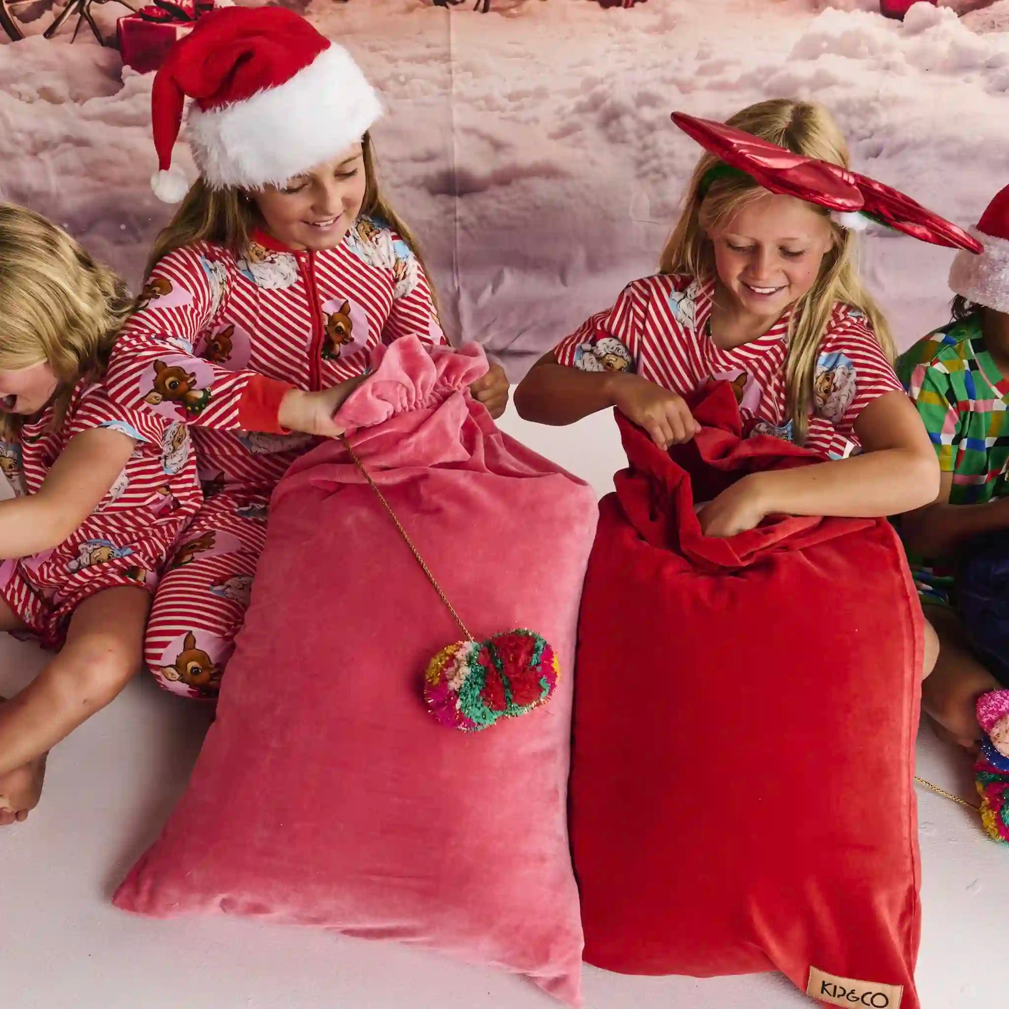 Two children in pajamas with Santa hats opening pink and red gift bags on a snowy background.