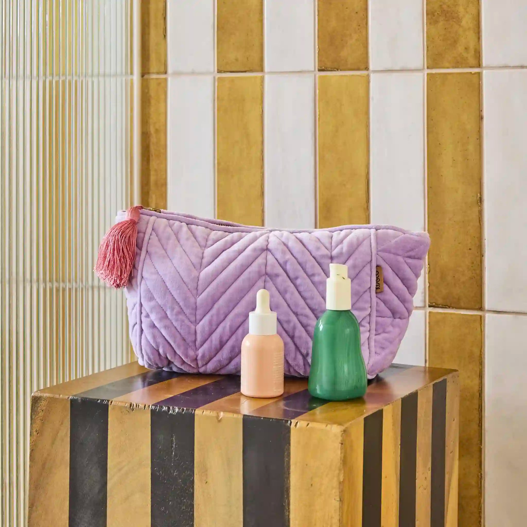 Purple quilted toiletry bag with tassels on a striped surface against a tiled wall.