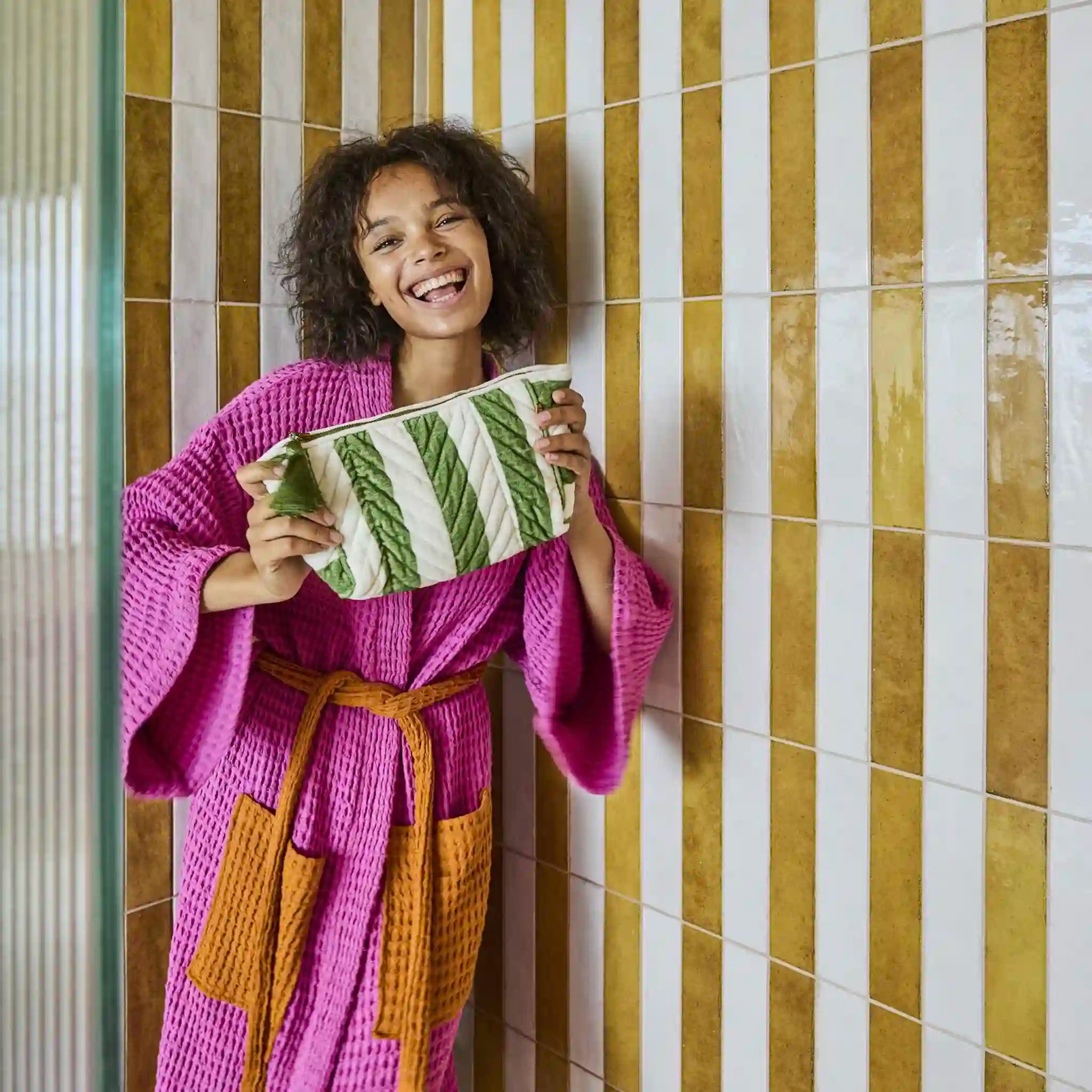 Woman in a pink robe holding a green and white striped toiletry bag against a striped tiled wall.