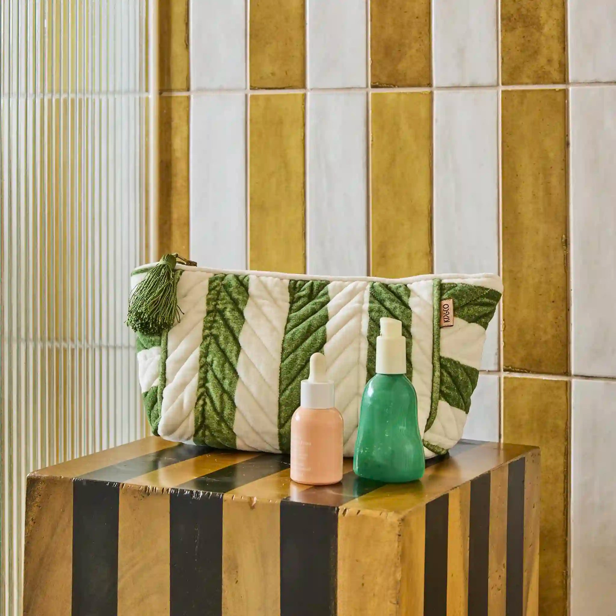Two bottles on a wooden block with a green and white patterned toiletry bag against a tiled wall.