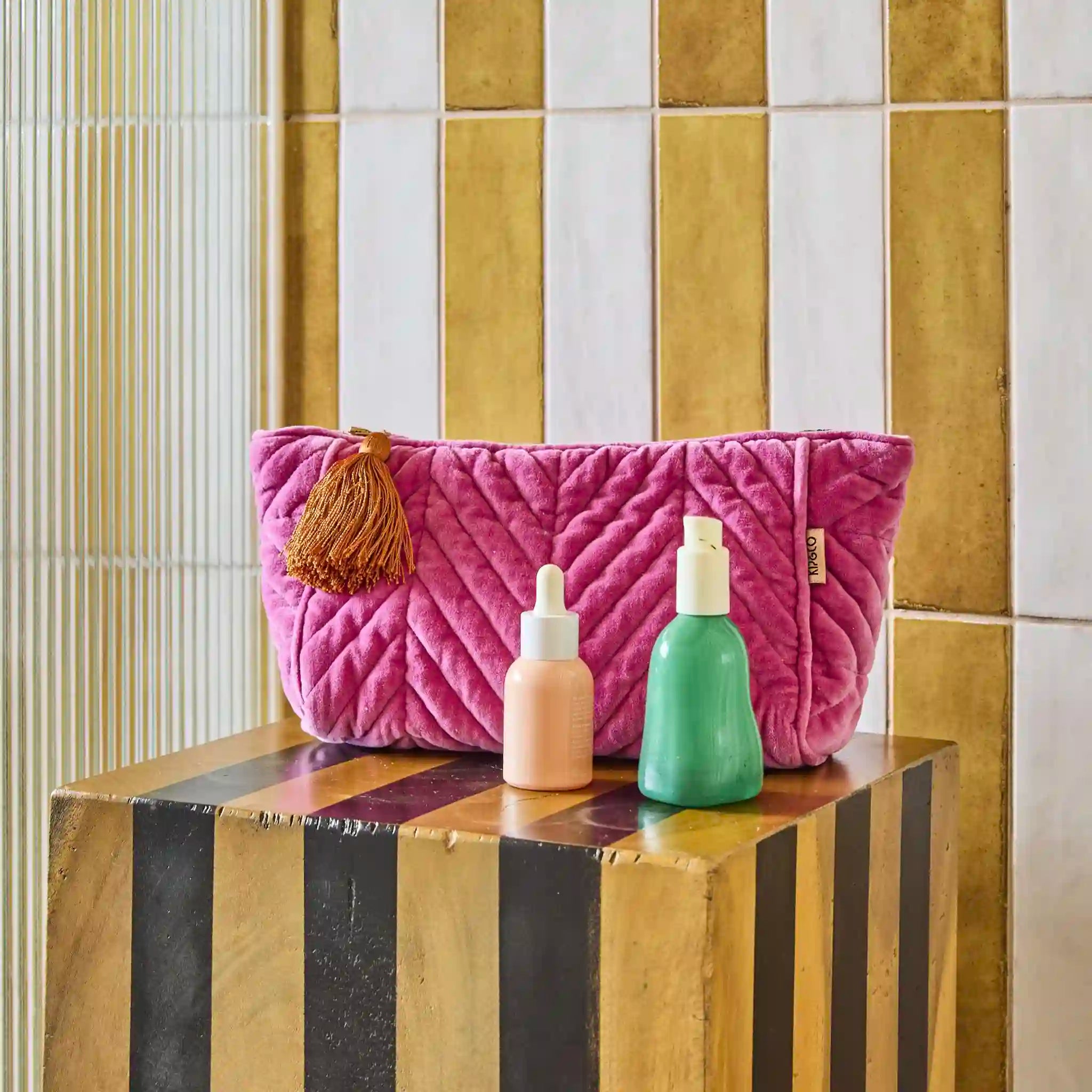Pink quilted toiletry bag with tassel on a striped surface, with two small bottles beside it.