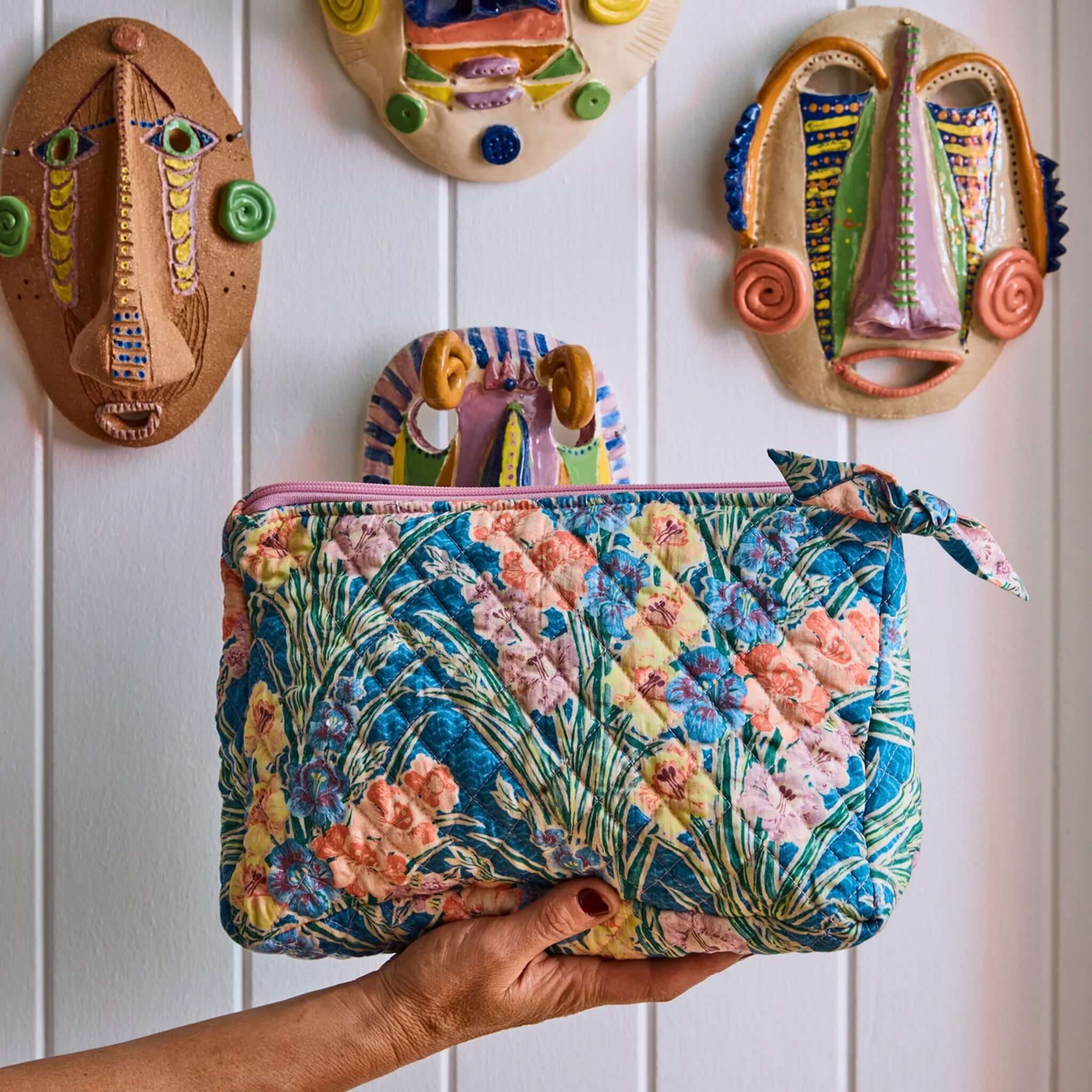 Kip & Co x Anna Spiro Quilted Toiletry Purse - Gladiolus held against colorful masks on a wall.