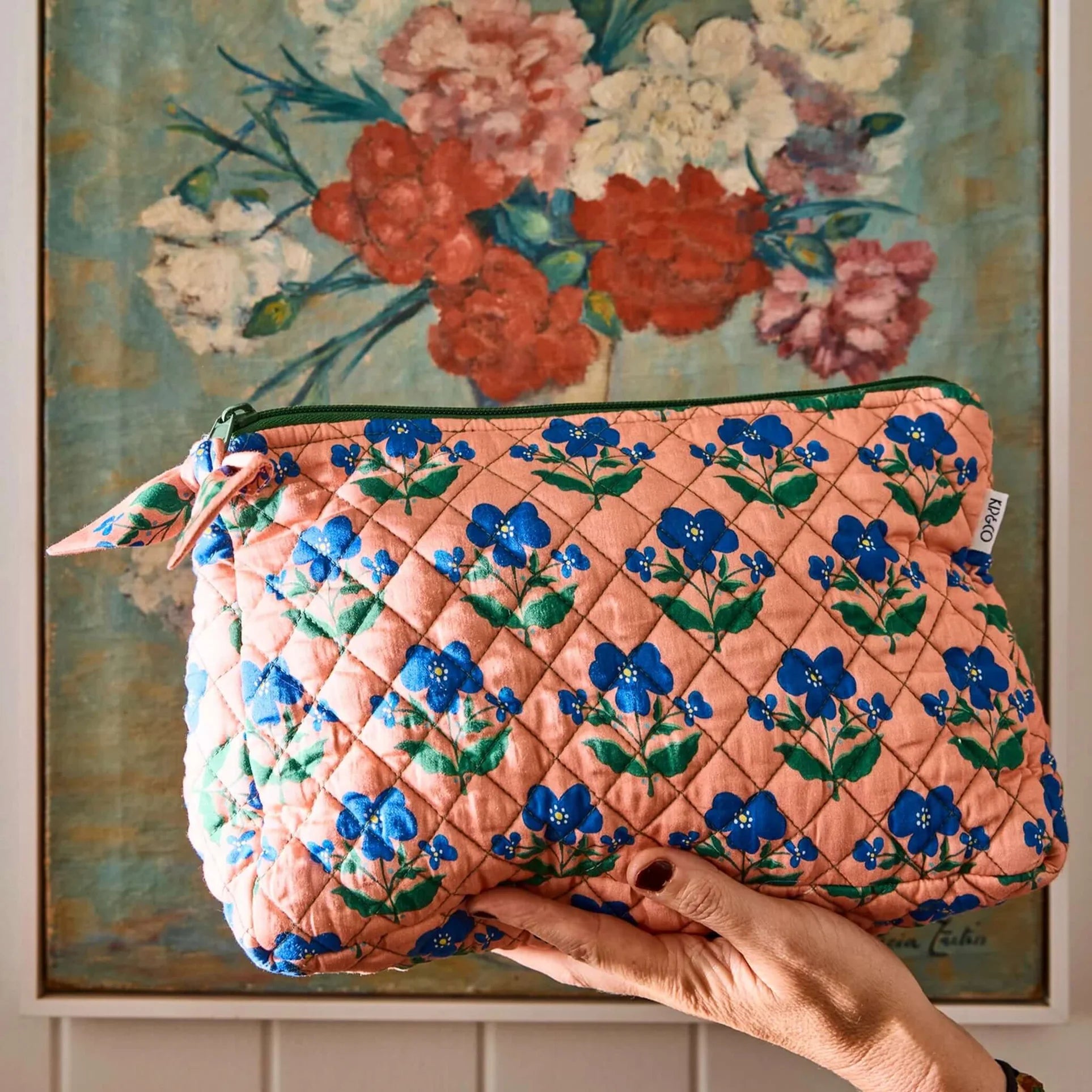 Floral-patterned quilted pouch held by a hand in front of a floral painting