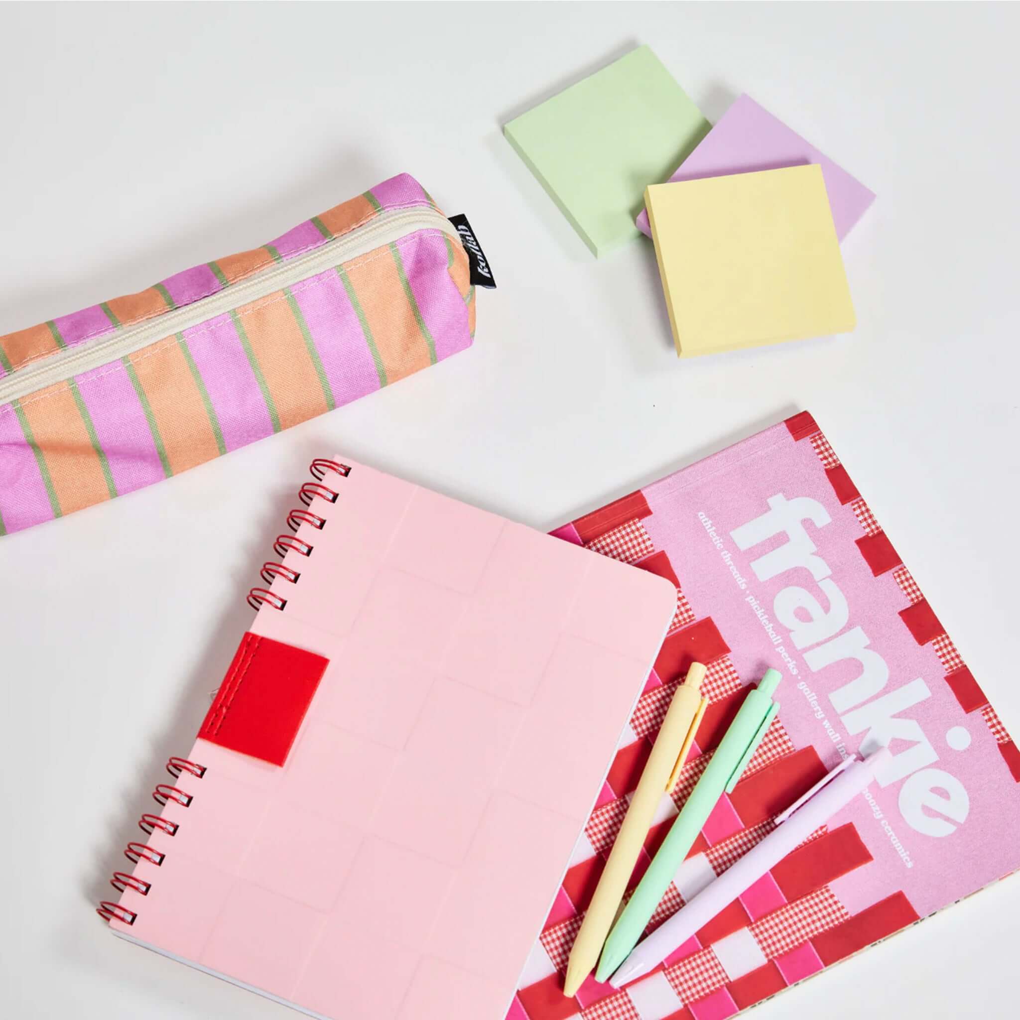 Stationery items including a pink notebook, striped pencil case, and colorful pens on a white background.