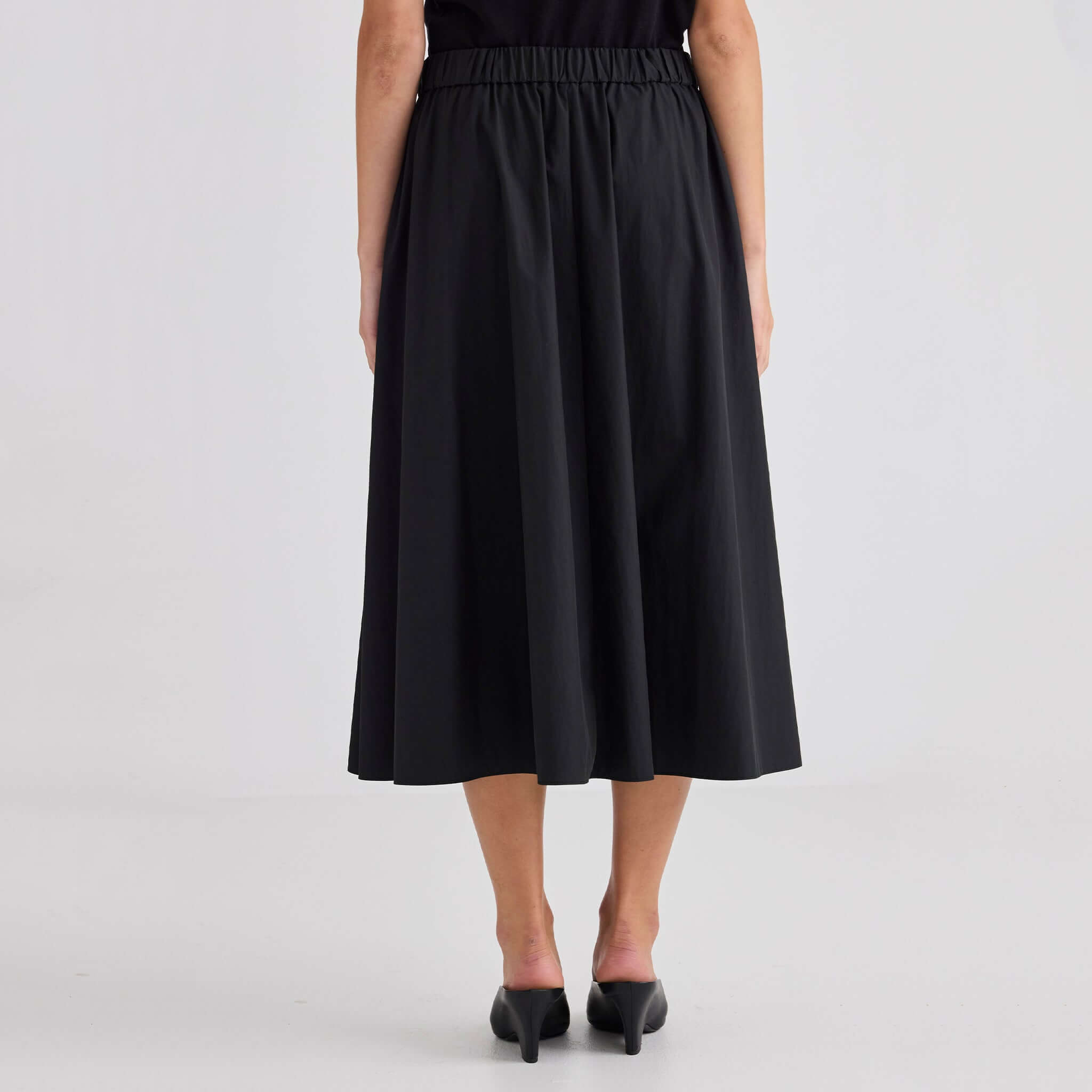 Rear view of a model wearing the Laing Ellis Pleated Skirt - Black, featuring an elasticated waistband and flowing pleats.