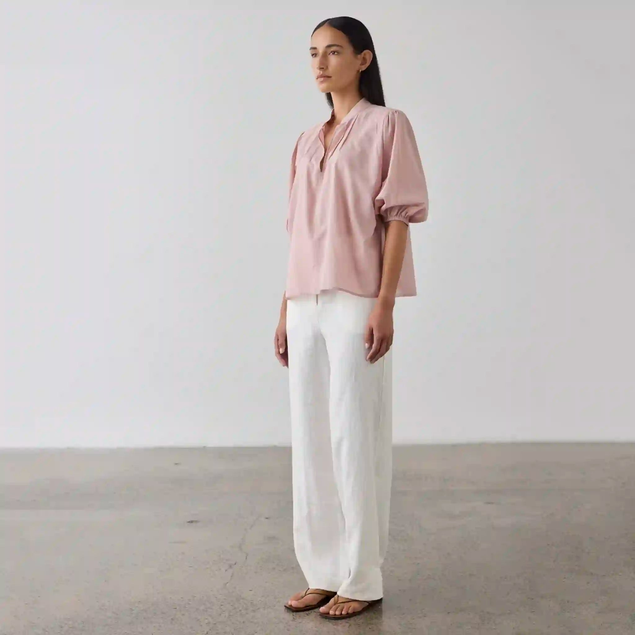 Woman wearing a pink blouse and white pants standing against a plain background