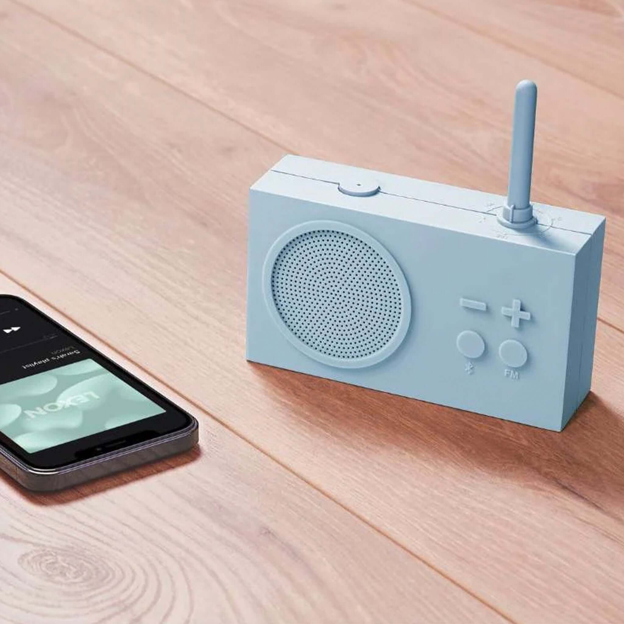 Lexon TYKHO 3 FM Radio & Bluetooth Speaker - Light Blue on a wooden table next to a smartphone.
