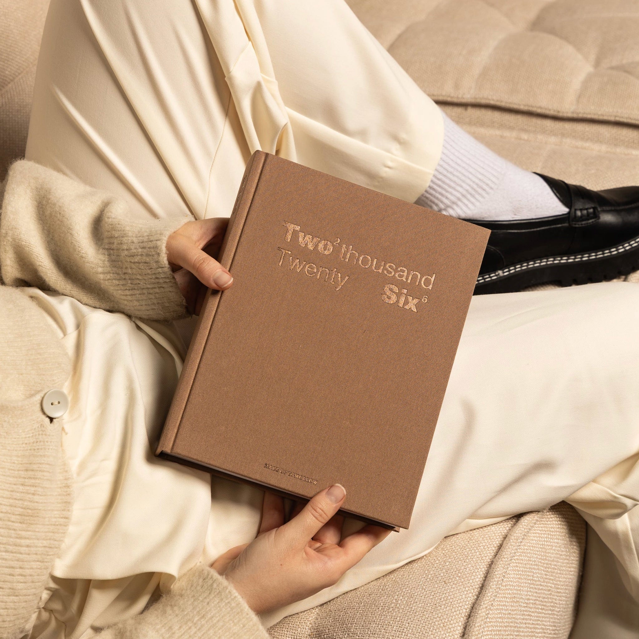 Brown book titled 'Two thousand Twenty Six' held by a person wearing a beige sweater.