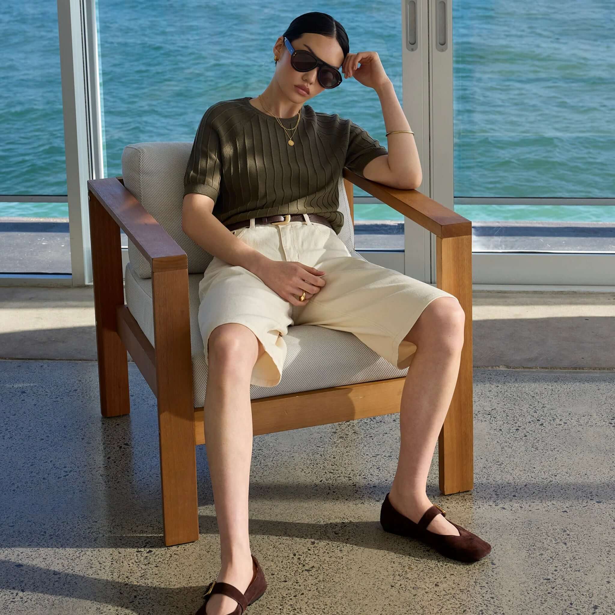 Woman wearing Marlow Soleil Knit Tee - Olive, sitting on a chair by the window with sea view.