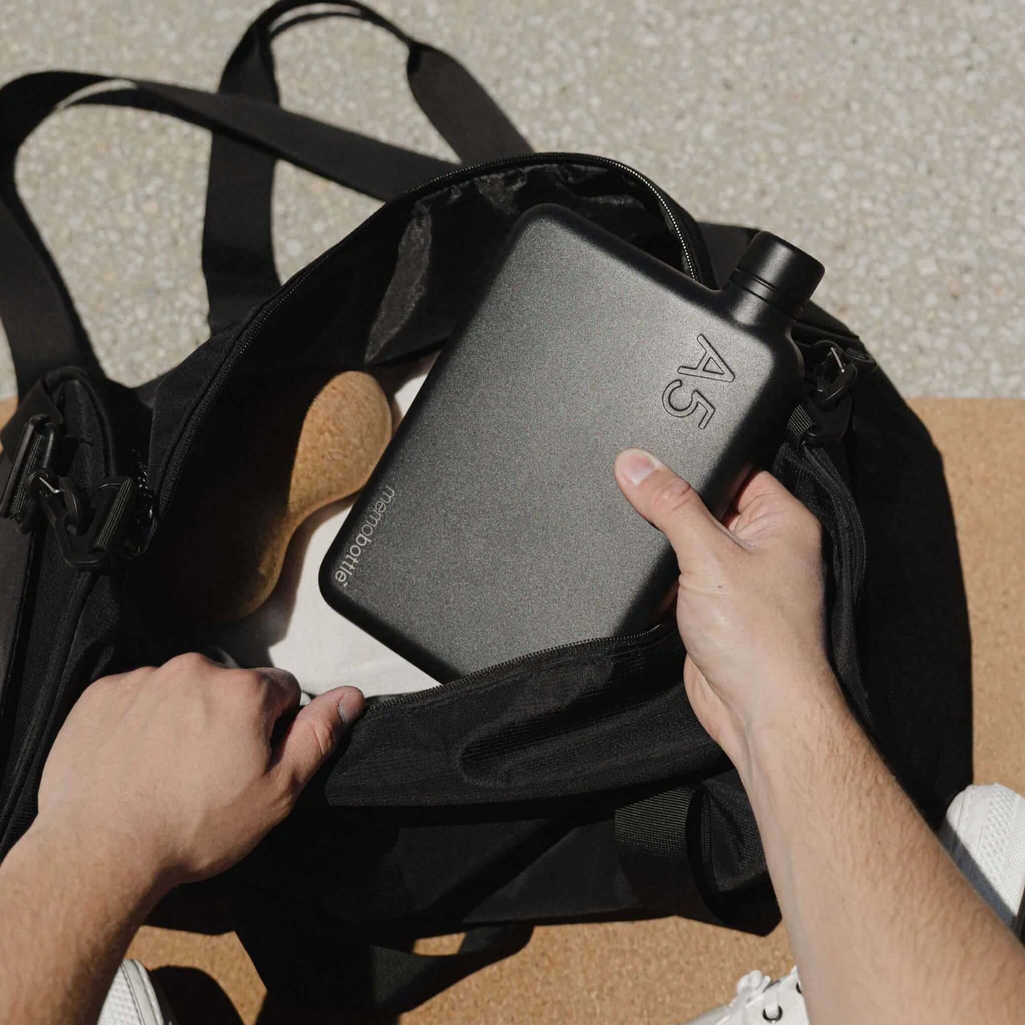 Alt Tag: Memobottle A5 Stainless Steel - Black flat water bottle being placed in a backpack, space-saving all-day hydration design