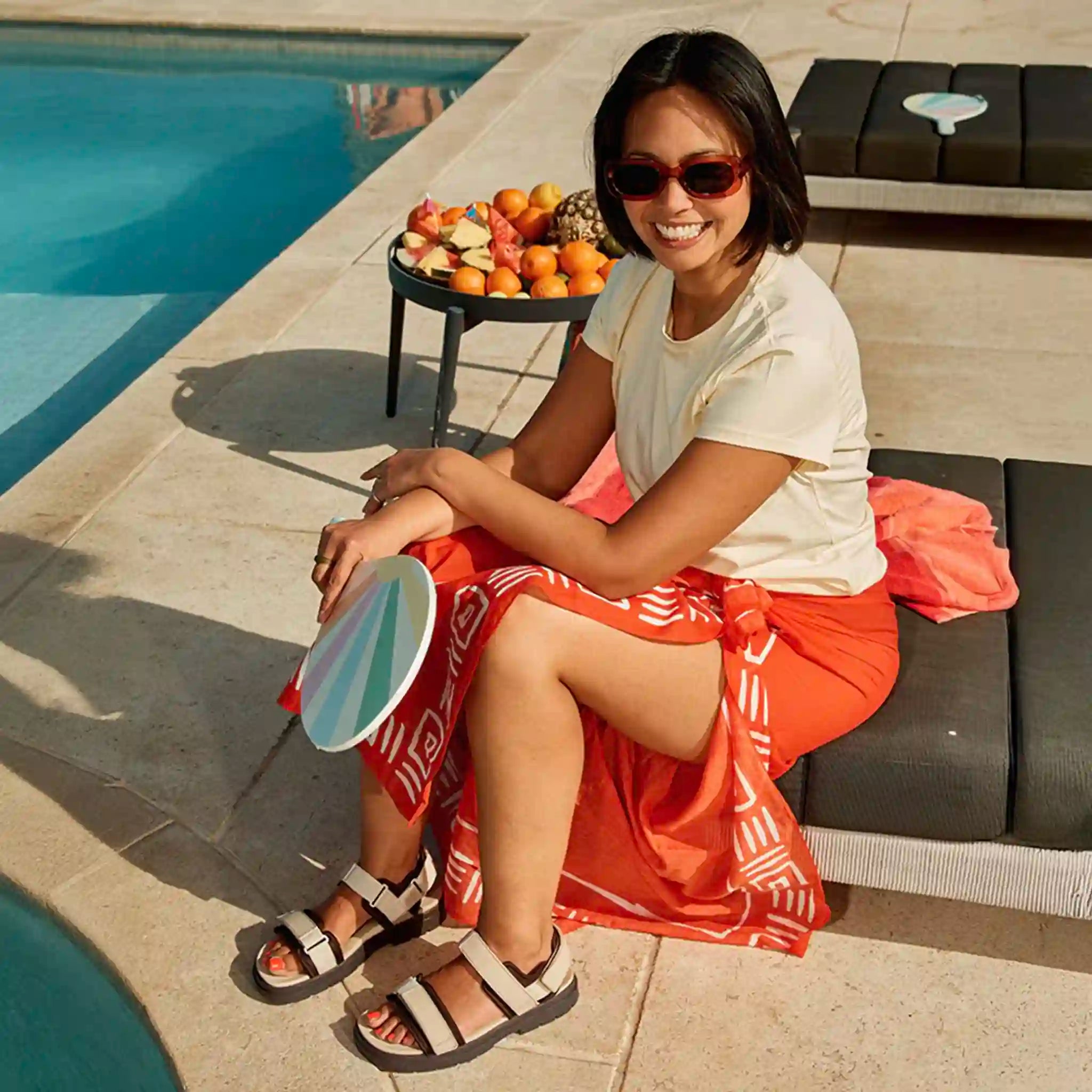 Woman sitting by a poolside with a fruit platter, wearing sunglasses and a white shirt with an orange skirt.