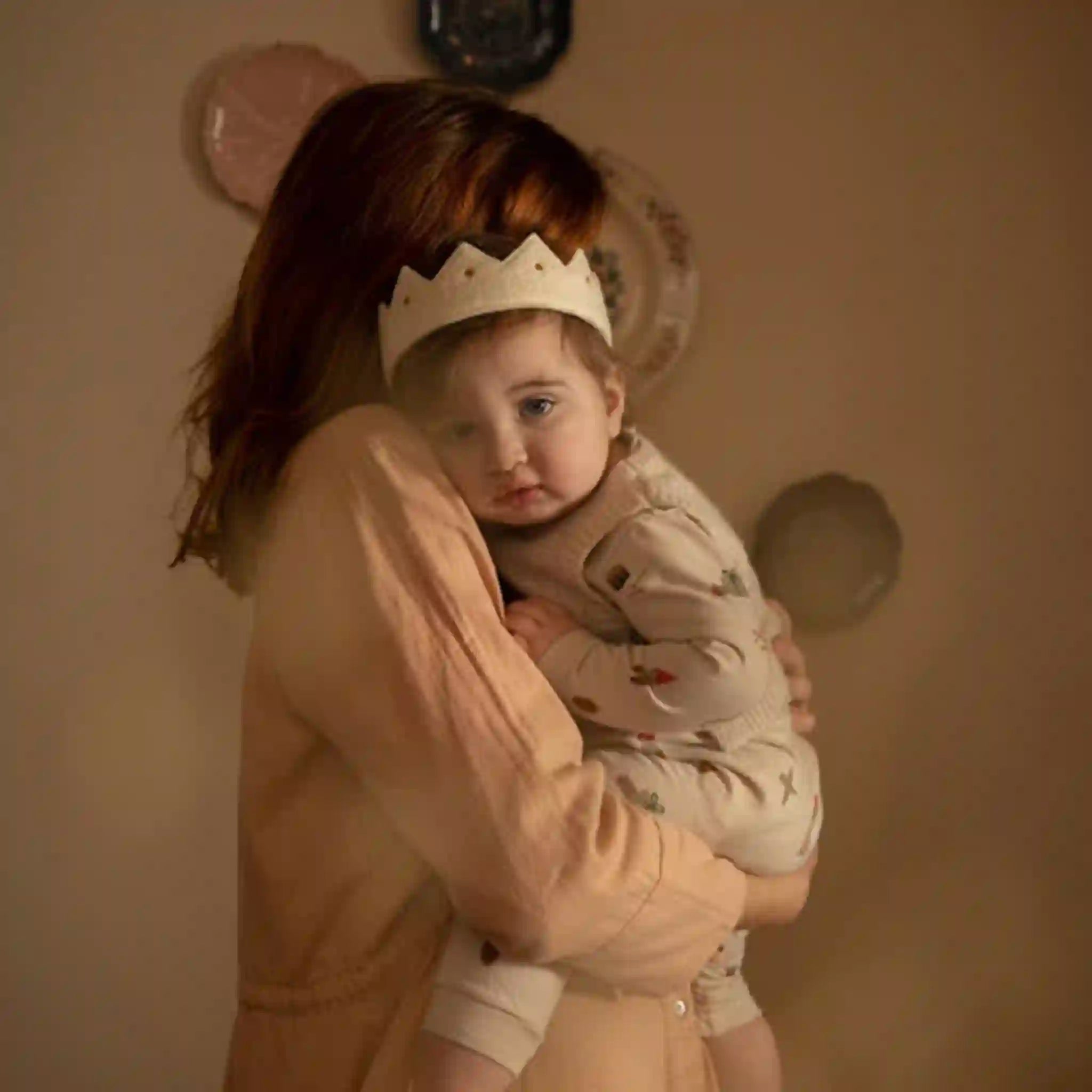 Woman holding a child wearing a white crown in a warm-toned room.