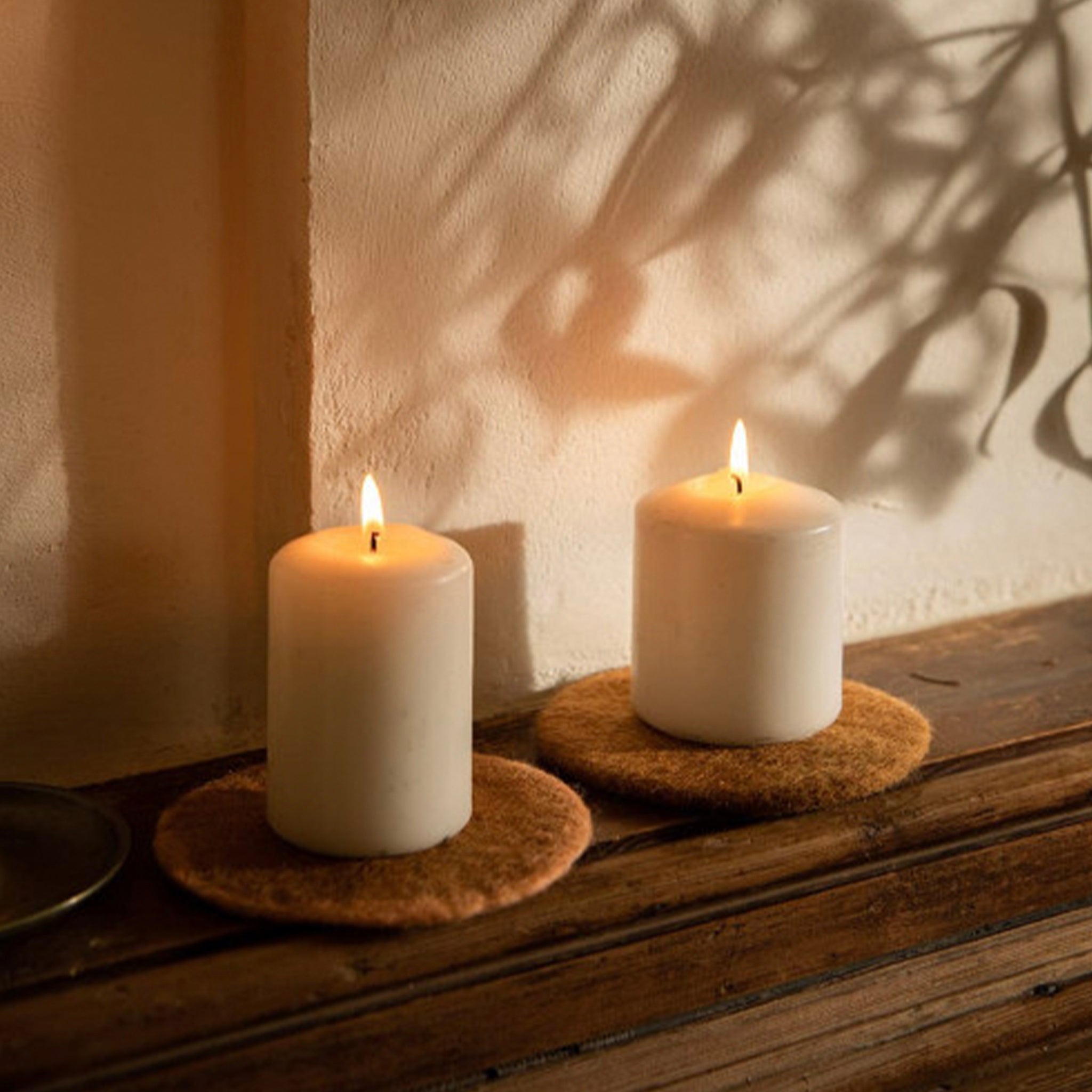 Two white candles on felt coasters with a warm glow against a neutral wall.