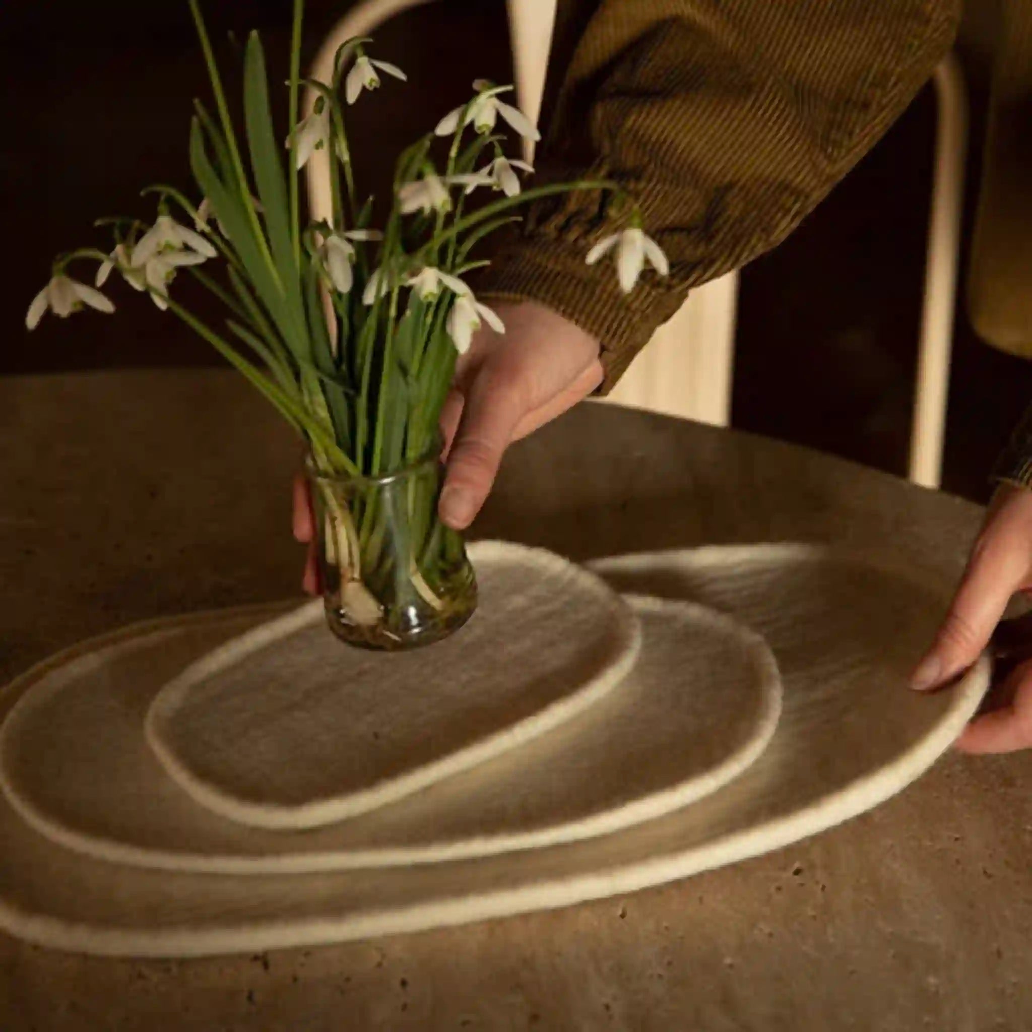 Person holding a glass vase with white flowers on a wooden surface