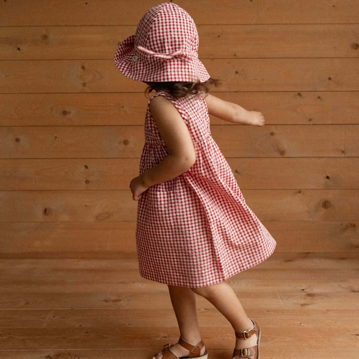 Nature Baby Organic Cotton Gingham Sunhat - Red Check on a child wearing a matching dress, playing indoors.