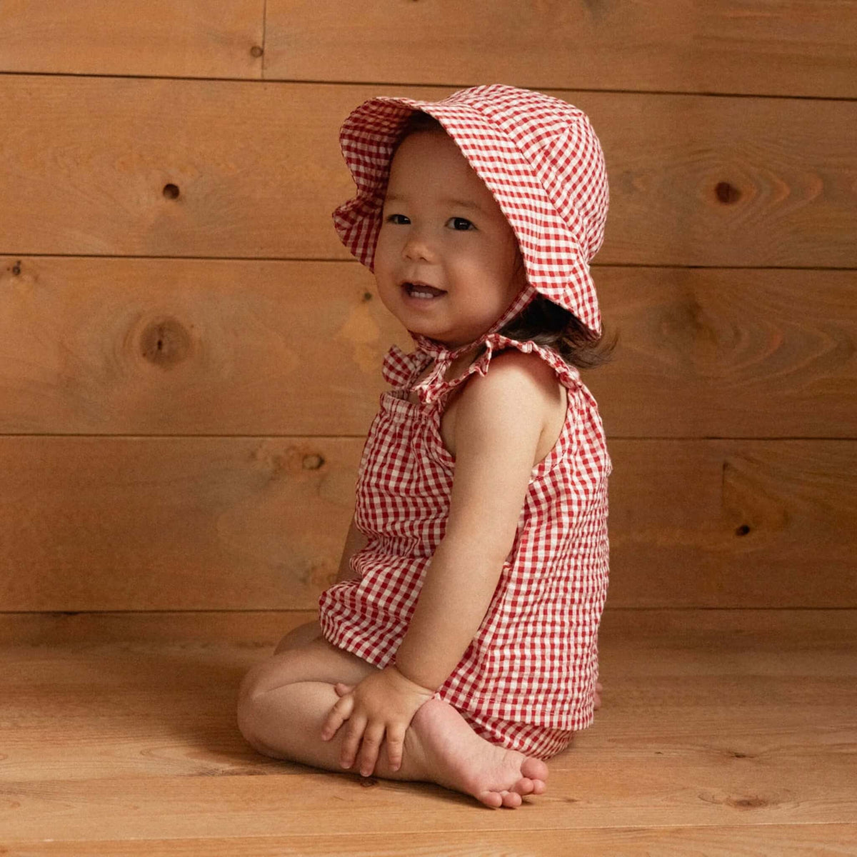 Baby wearing Nature Baby Organic Cotton Gingham Sunhat - Red Check sitting on wooden floor, smiling with a playful look.