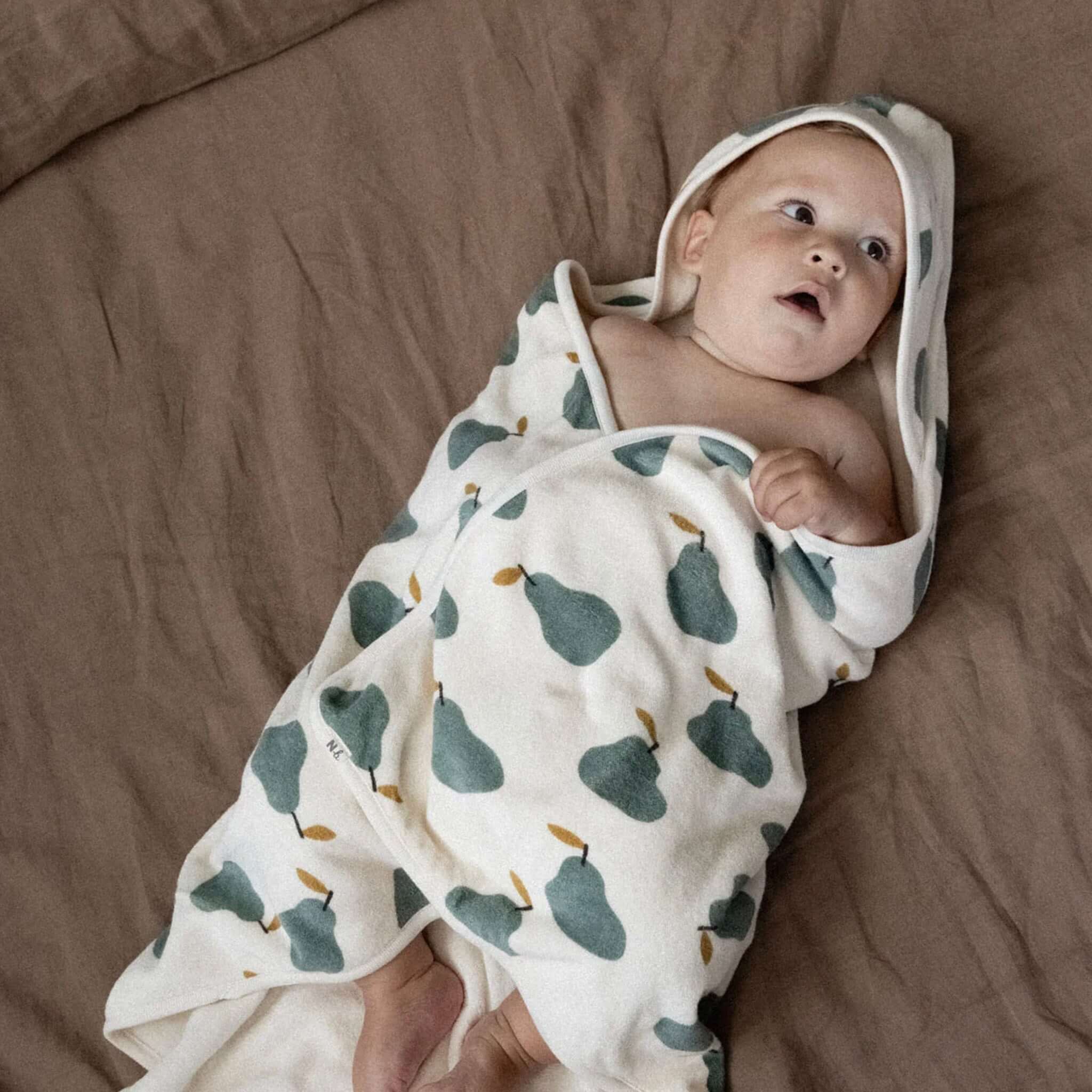 Baby wrapped in Nature Baby Organic Cotton Hooded Towel - Pear on a soft bed, showcasing the cozy design and bear ears.